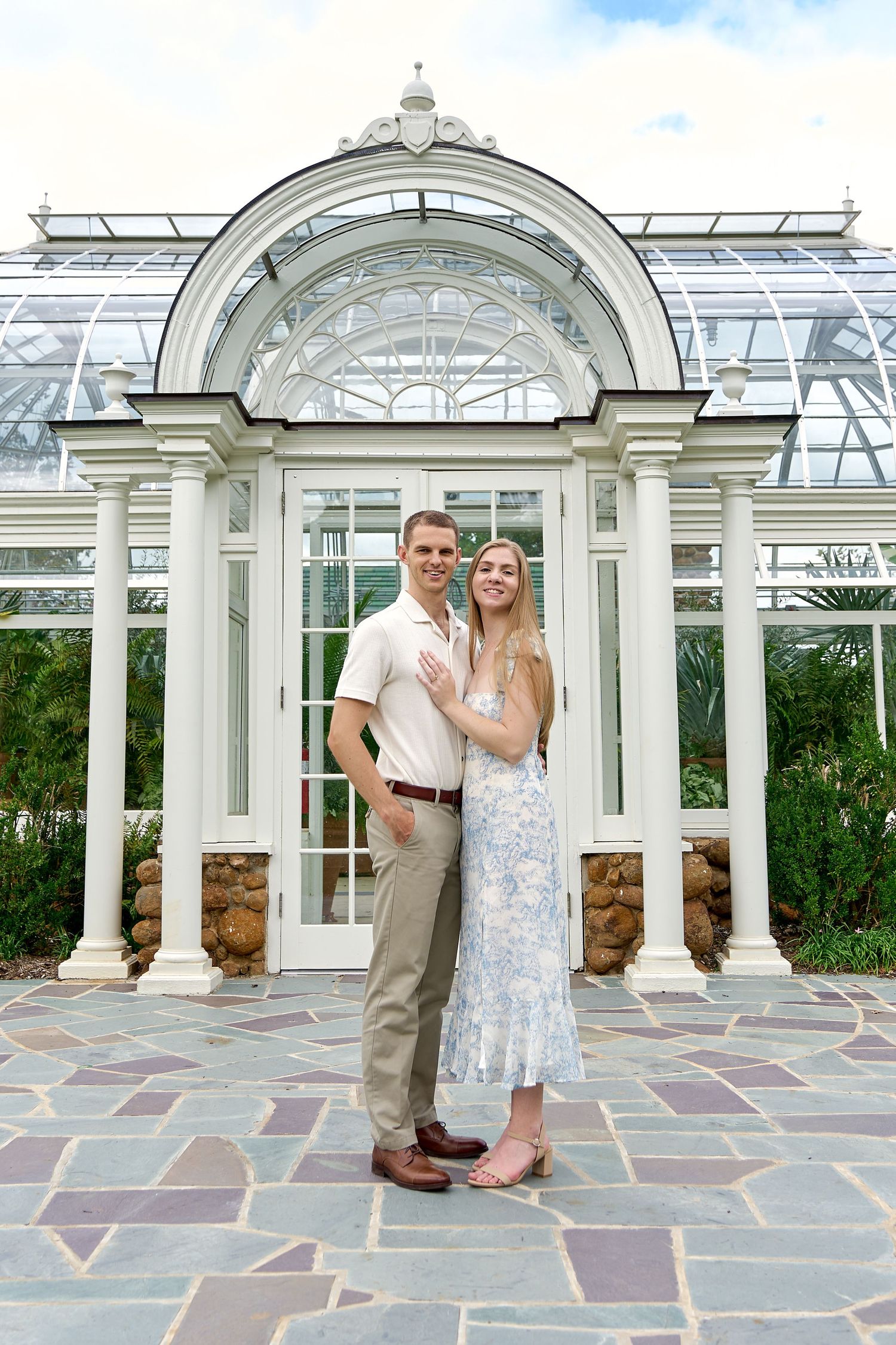 Smiling couple posing in front of ornate glass greenhouse on stone patio.