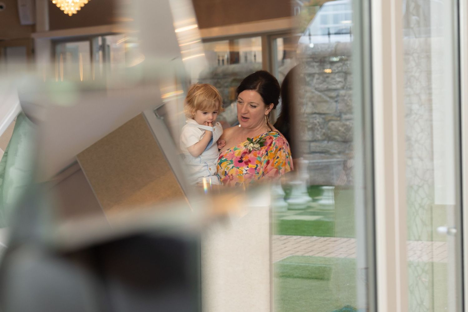 Woman in floral dress holding a toddler, photographed through a glass window at a wedding venue.