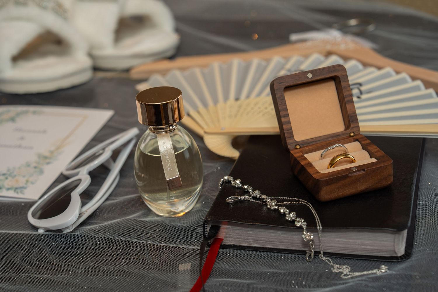Wedding flat lay with perfume bottle, ring box with gold band, bracelet, hand fan, and white heels on gray surface.
