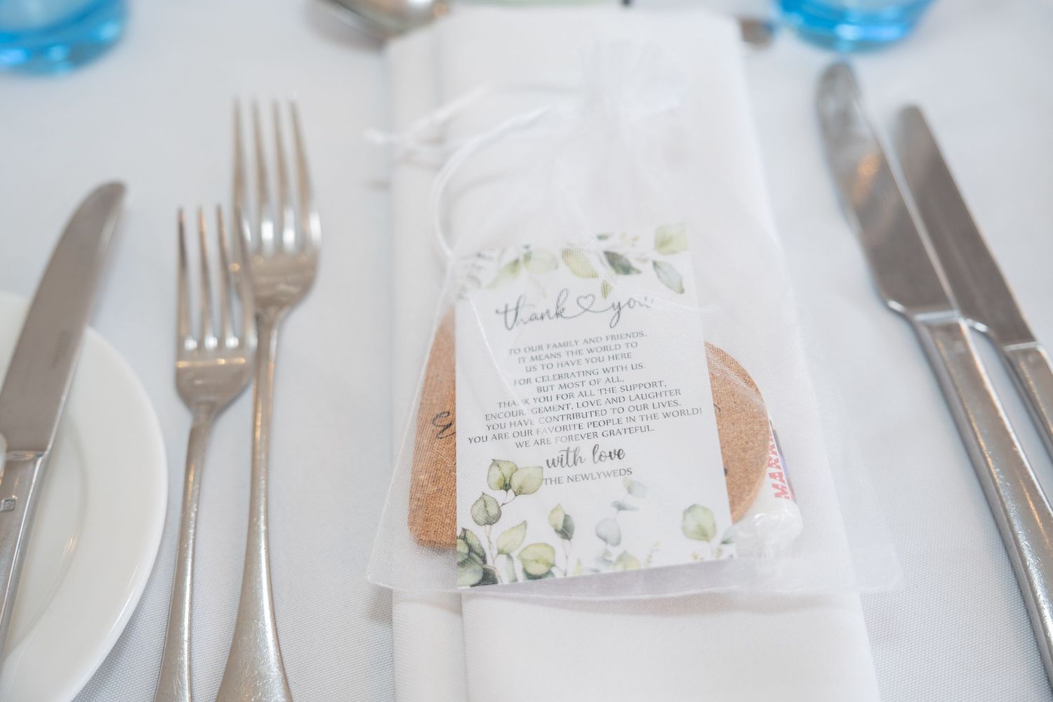 Elegant wedding table setting with silverware, white napkin, and a floral thank you card favor on a white tablecloth.