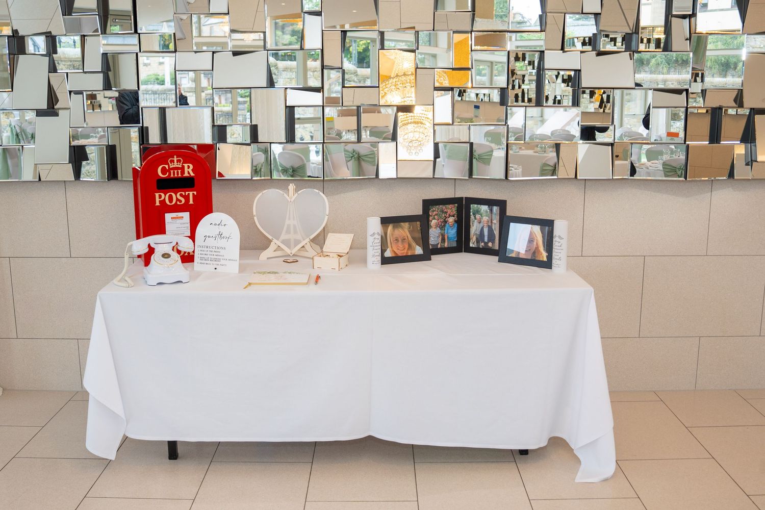 Elegant wedding gift table with white tablecloth, heart decor, photo frames, guest book, and red post box against mirror wall.