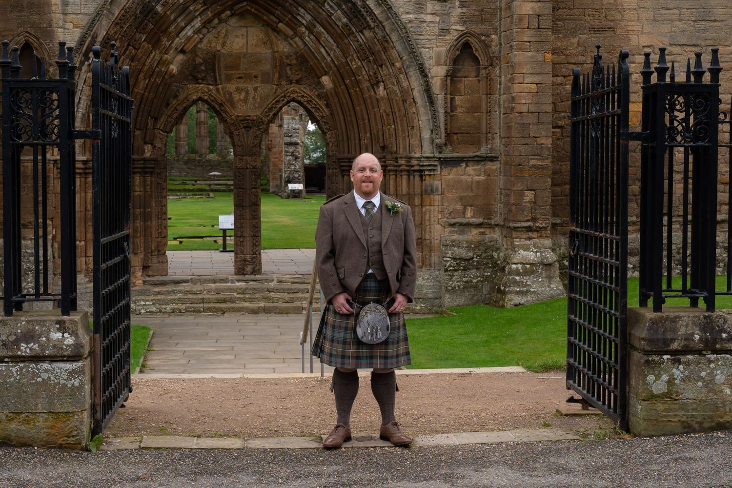 Man in traditional Scottish kilt and tweed jacket standing at historic abbey gate.