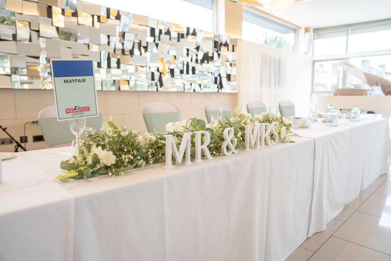 Elegant wedding head table with white linen, floral greenery, and decorative MR & MRS letters at a reception venue.