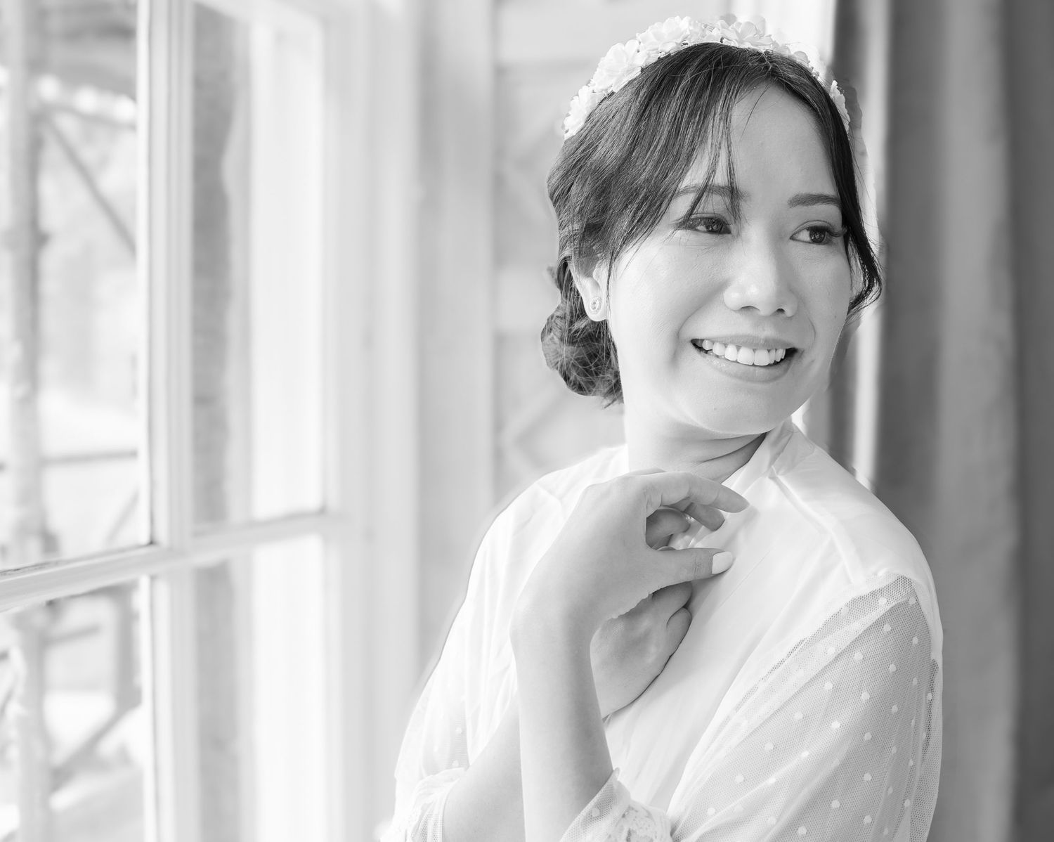 Beautiful bride in white dress smiling by a window in elegant black and white portrait photo.