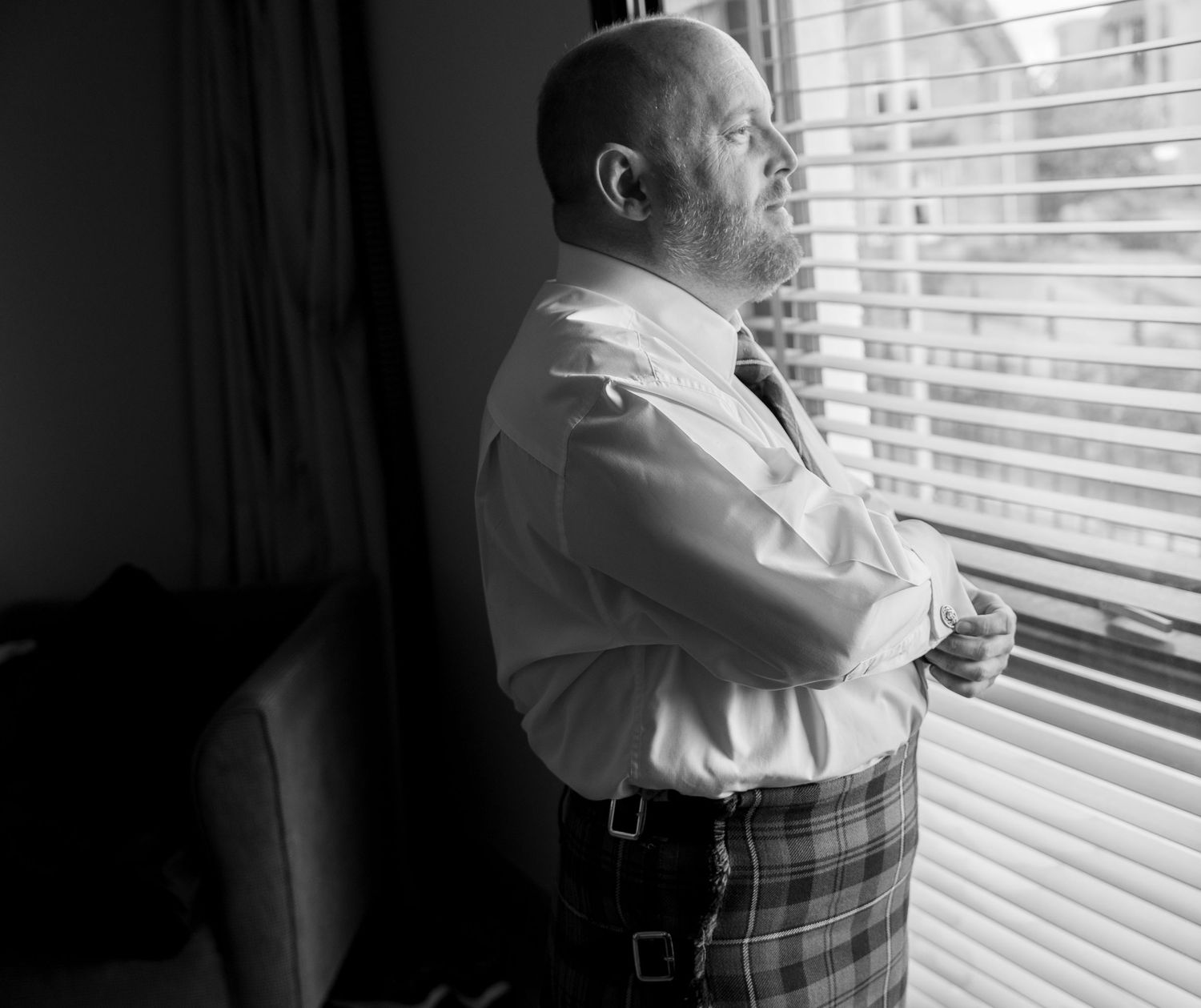 Bearded man in white shirt and tartan kilt standing by window with arms crossed in black and white photo.