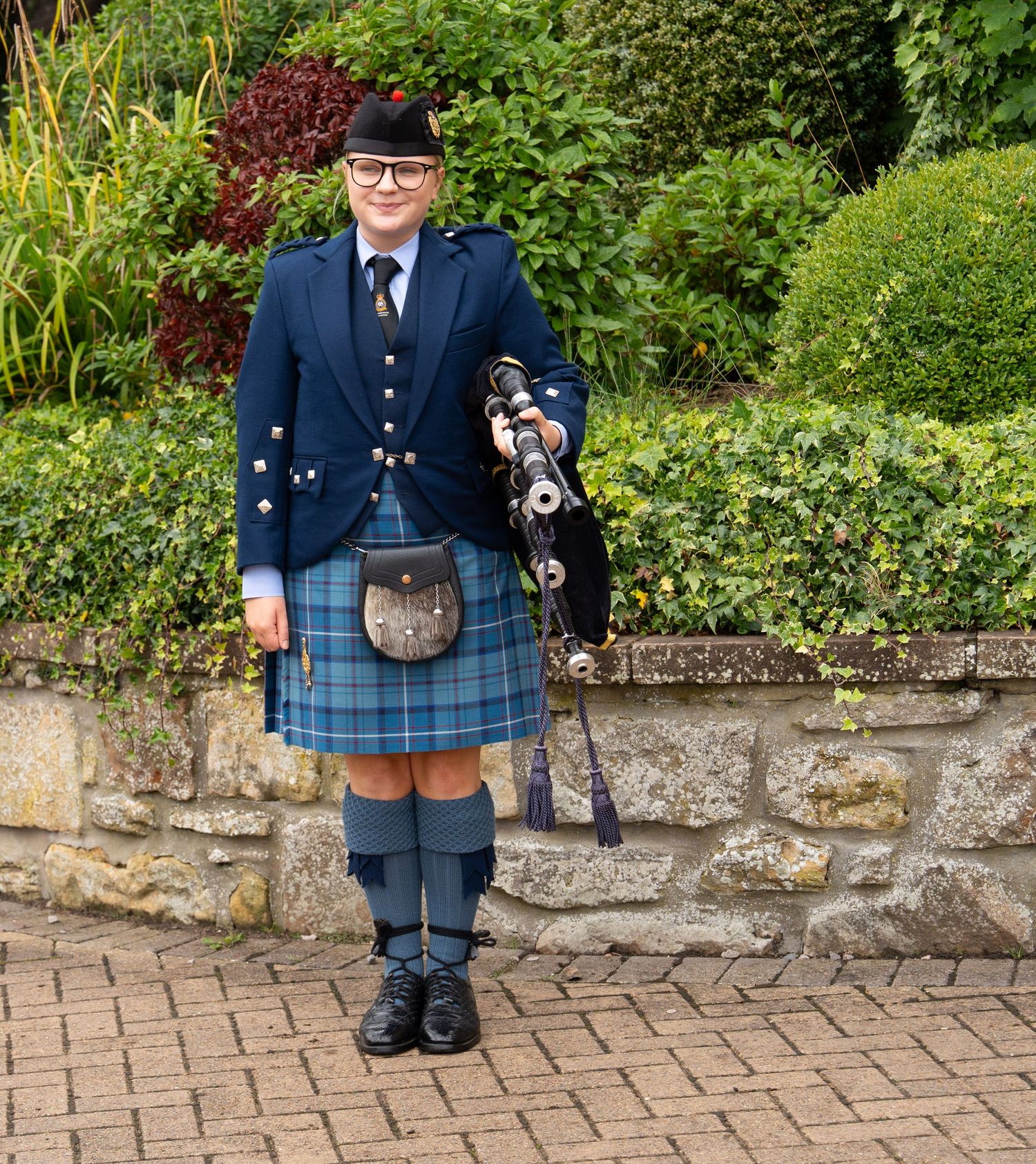 A bagpiper in traditional Scottish attire with blue jacket and tartan kilt stands in a garden.