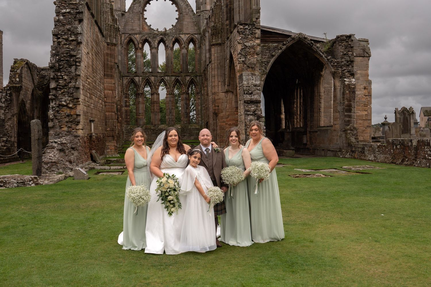 Wedding party posing in front of historic stone abbey ruins with bride, groom, bridesmaids in sage green gowns.
