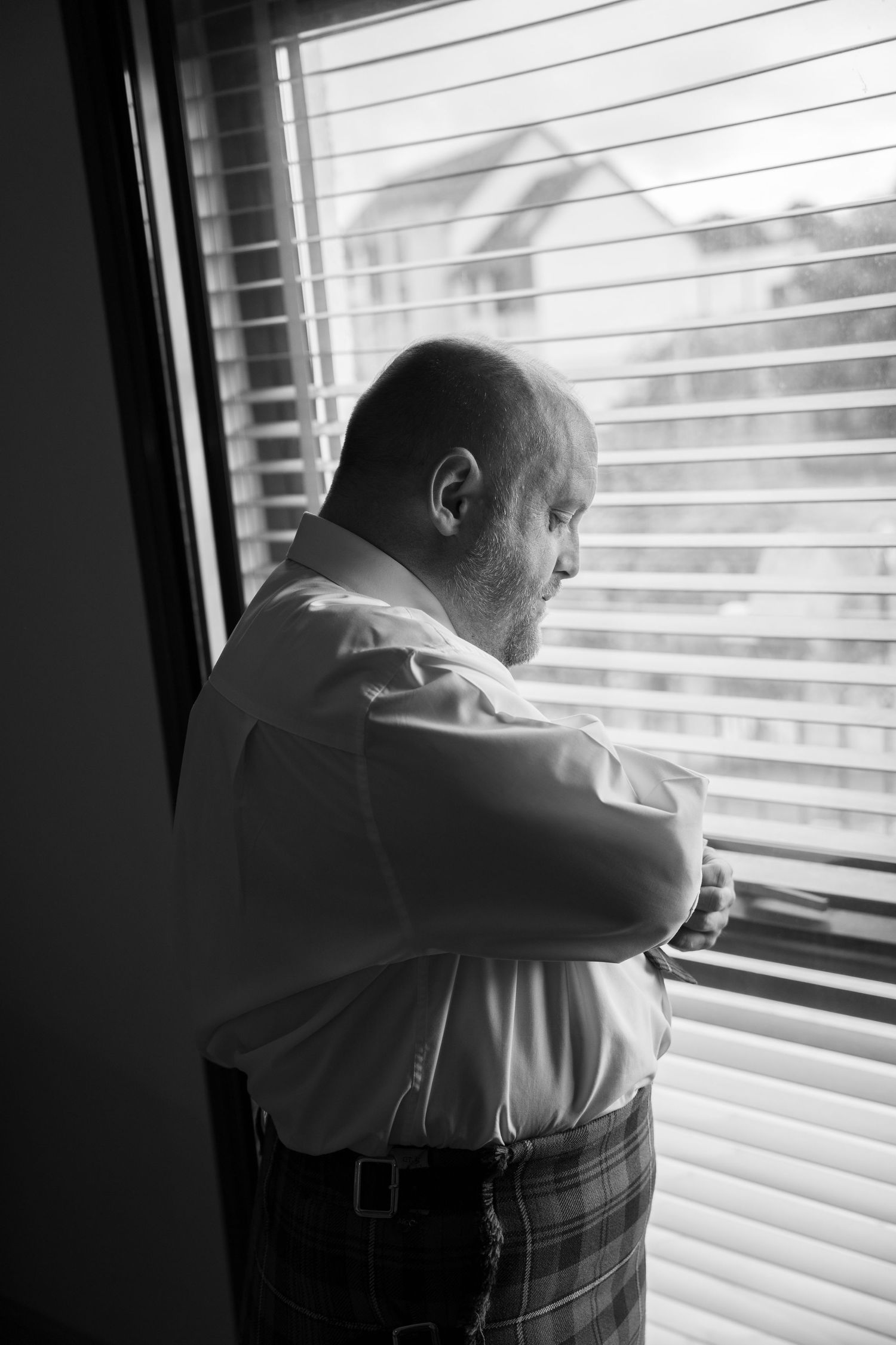 Black and white photo of groom in kilt adjusting cufflinks by window with venetian blinds.