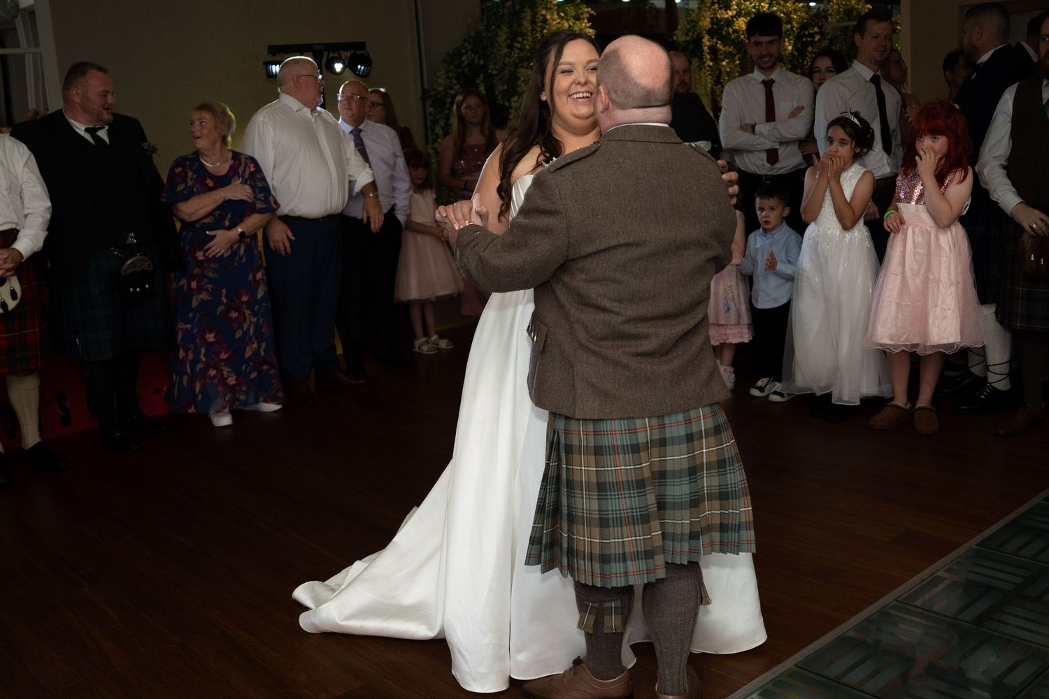 Bride in white gown dancing with a man in traditional Scottish tartan kilt surrounded by wedding guests on dance floor.
