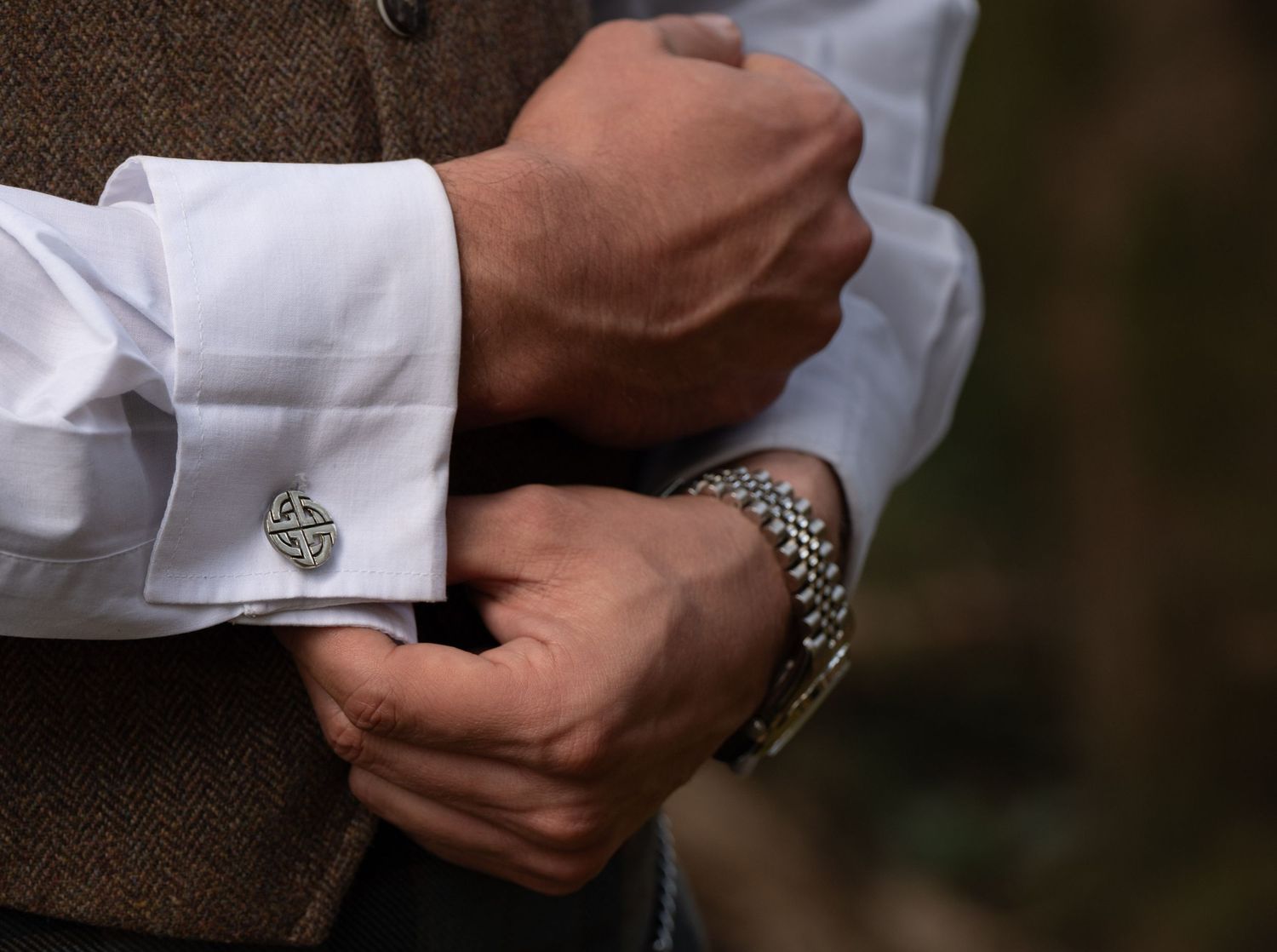 Man adjusting silver Celtic cufflinks on white dress shirt, wearing brown tweed vest and silver bracelet.