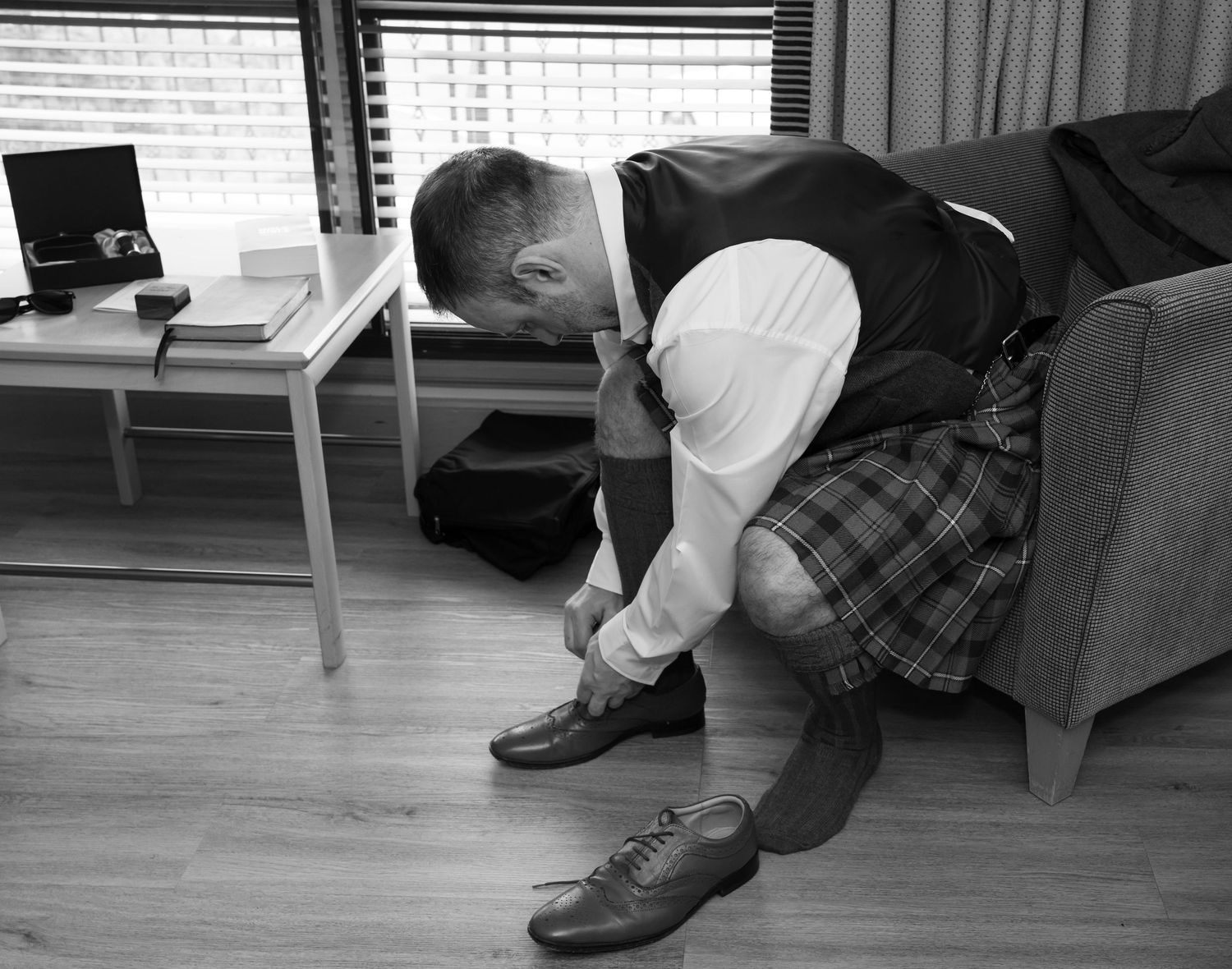 Man in Scottish kilt and waistcoat putting on dress shoes while seated on a sofa in a hotel room.