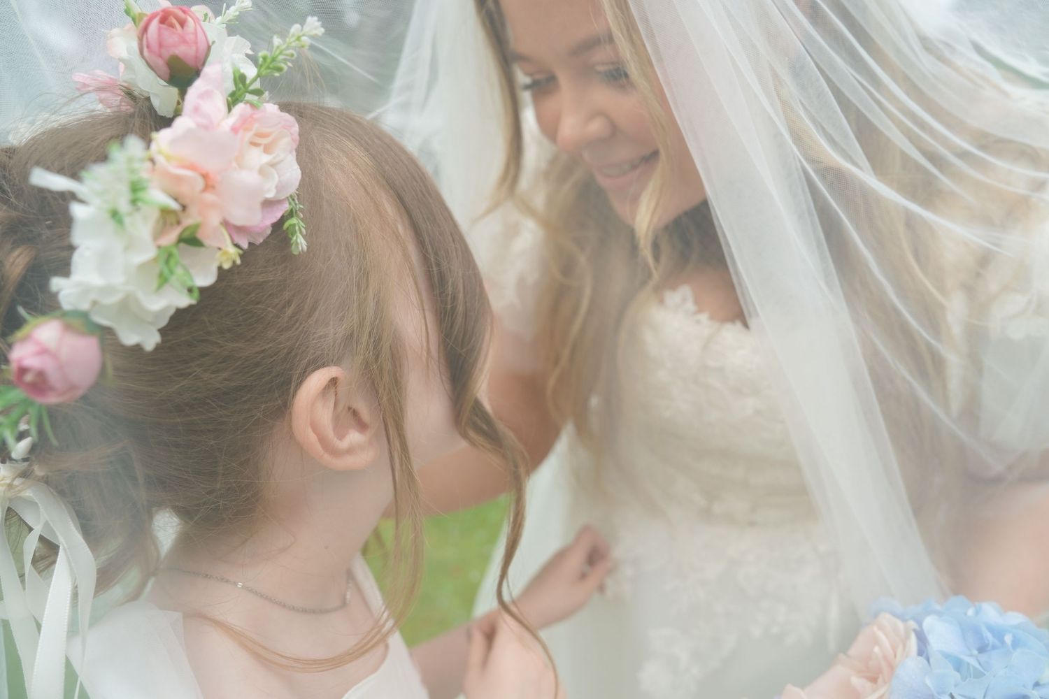 Bride in white lace gown shares a tender moment with flower girl wearing floral crown under veil.