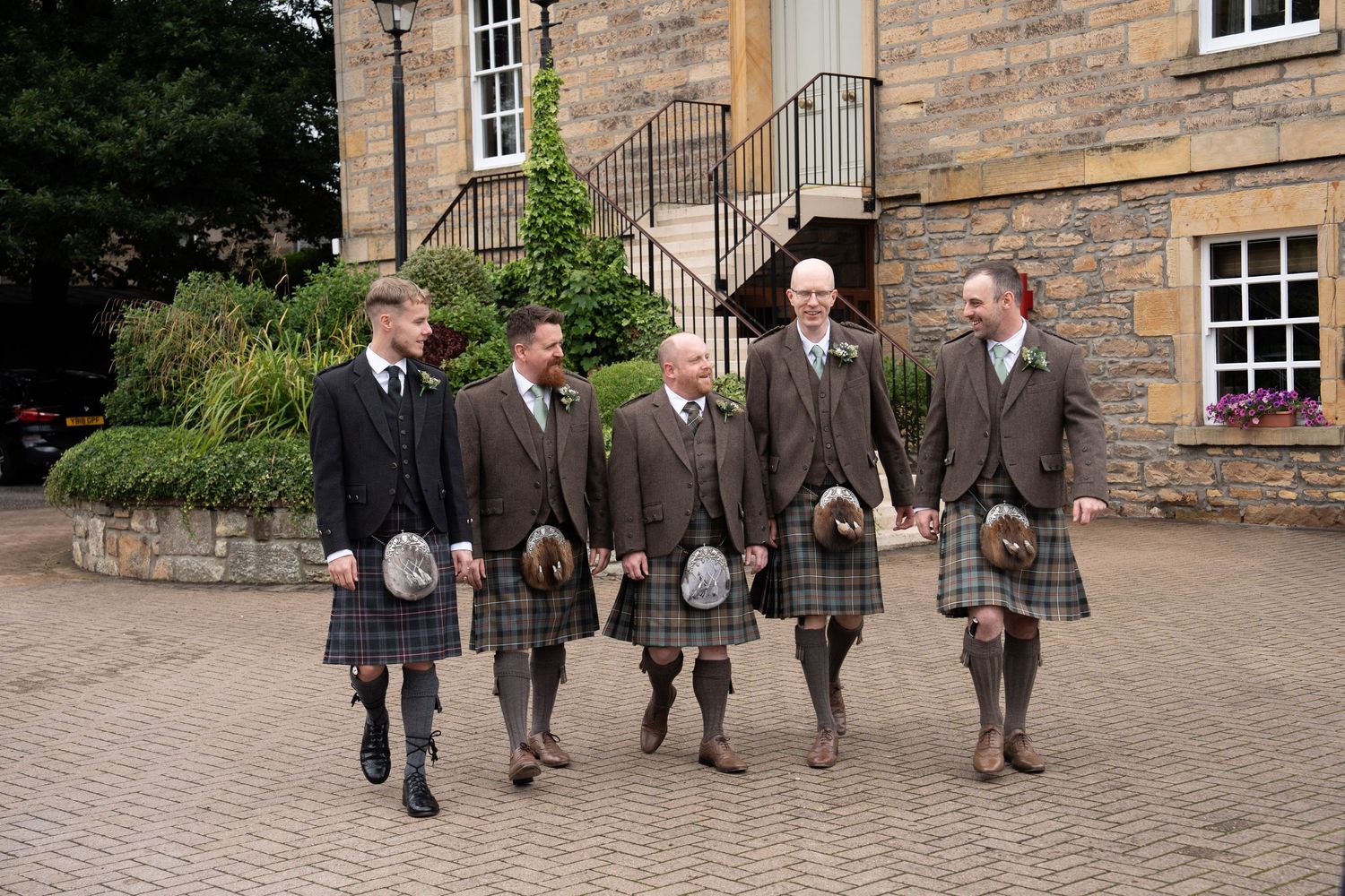 Five groomsmen in traditional Scottish kilts walking together outside a stone building.