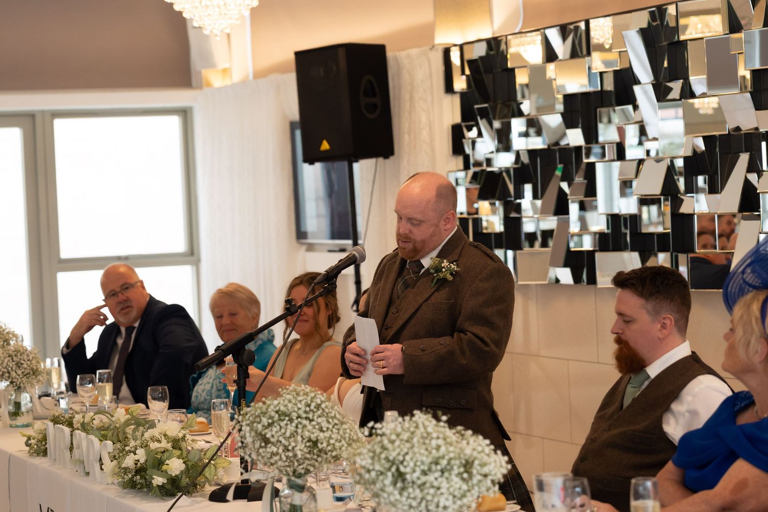 Man in olive suit giving a speech at a wedding reception with floral arrangements and guests seated at the head table.