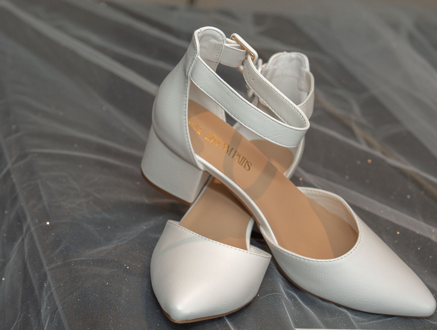 White block heel pointed toe pumps with ankle strap buckle closure, displayed on gray surface.