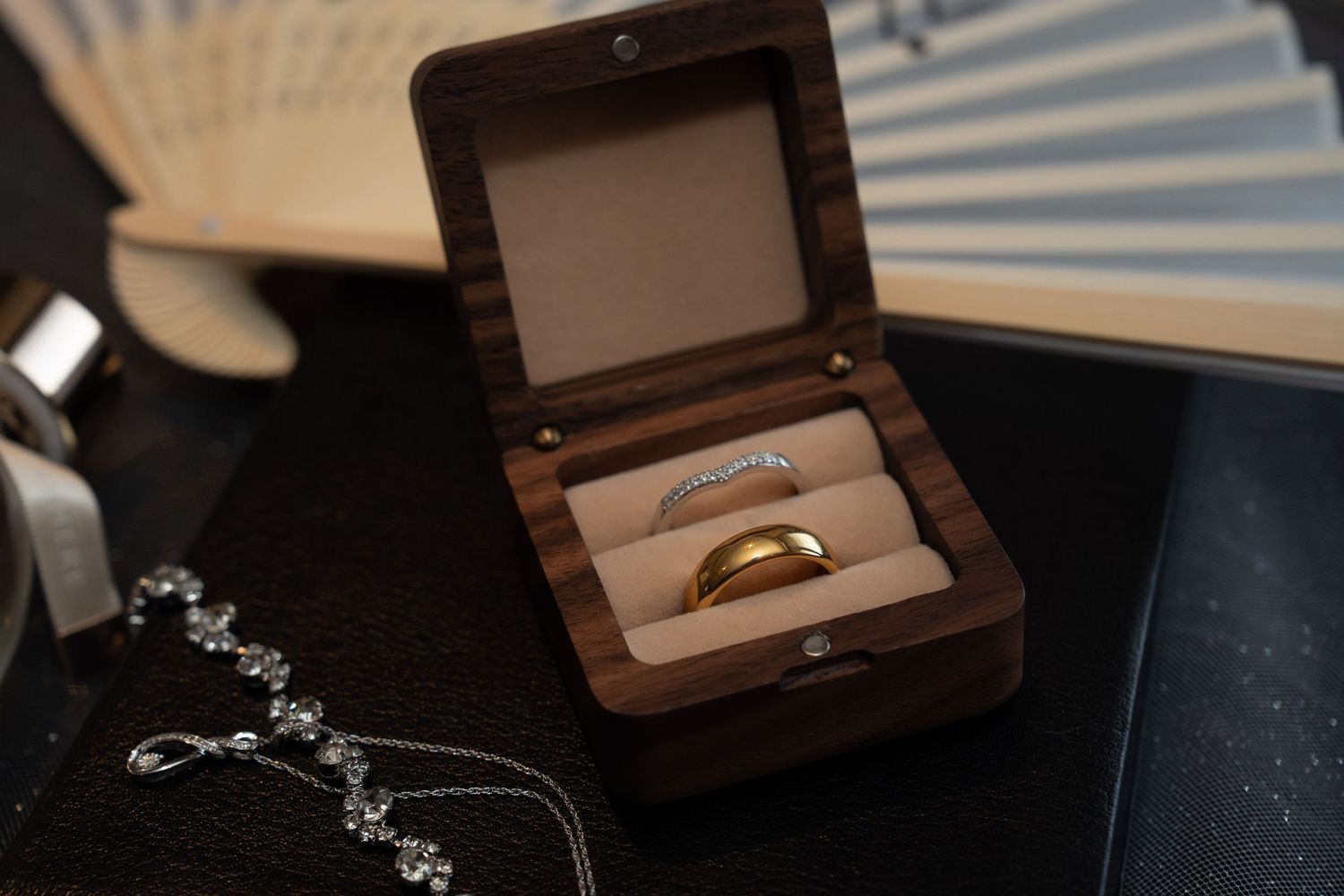 Two wedding rings, a gold band and diamond ring, displayed in a wooden ring box on a dark surface.