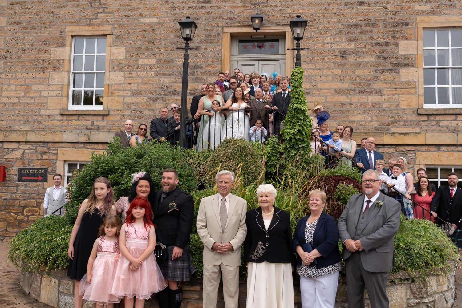 Large wedding group photo of guests and wedding party posing on steps outside a Scottish stone building.