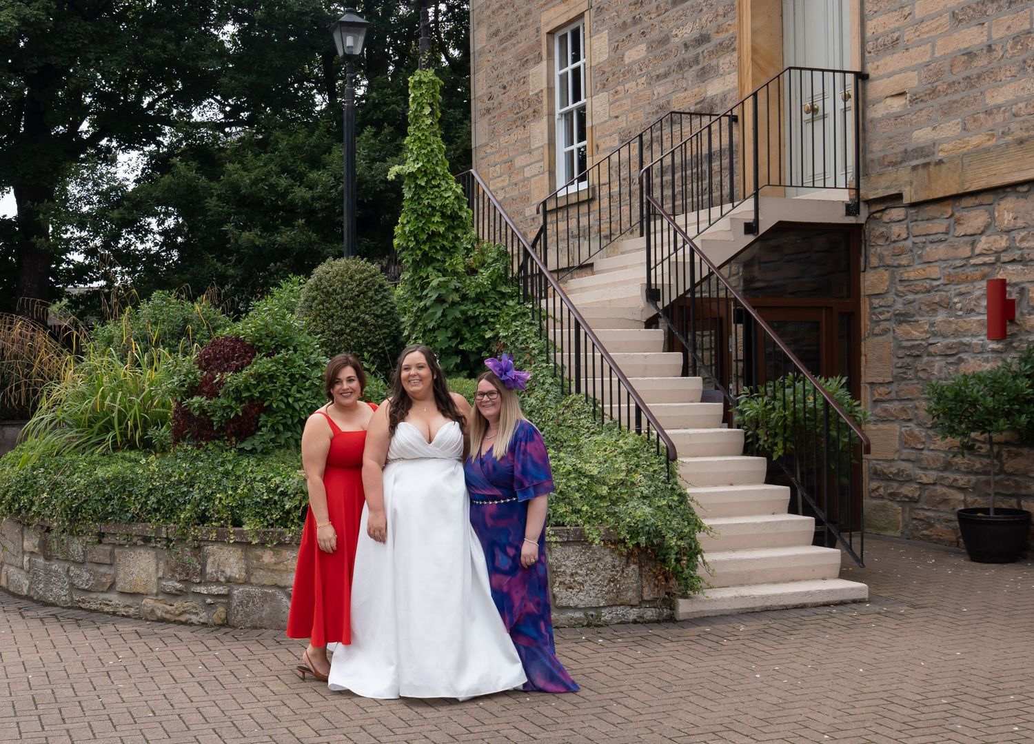 Bride in white gown poses with two guests outside elegant stone venue with staircase and lush greenery.