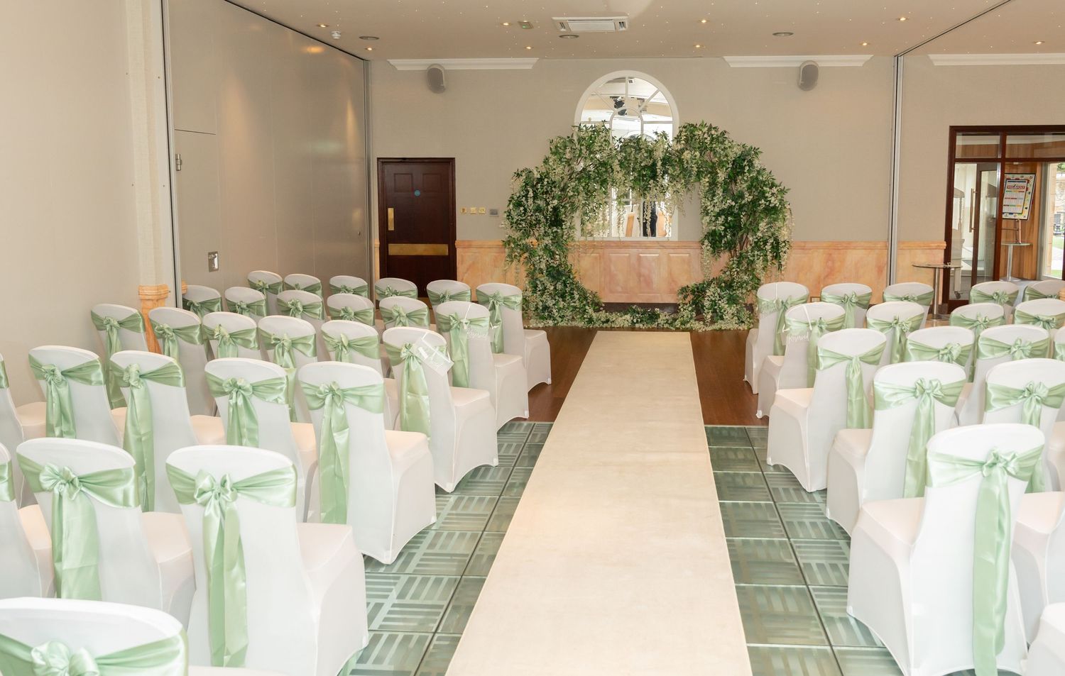 Elegant wedding ceremony room with white chair covers, sage green sashes, and a lush greenery arch at the altar.