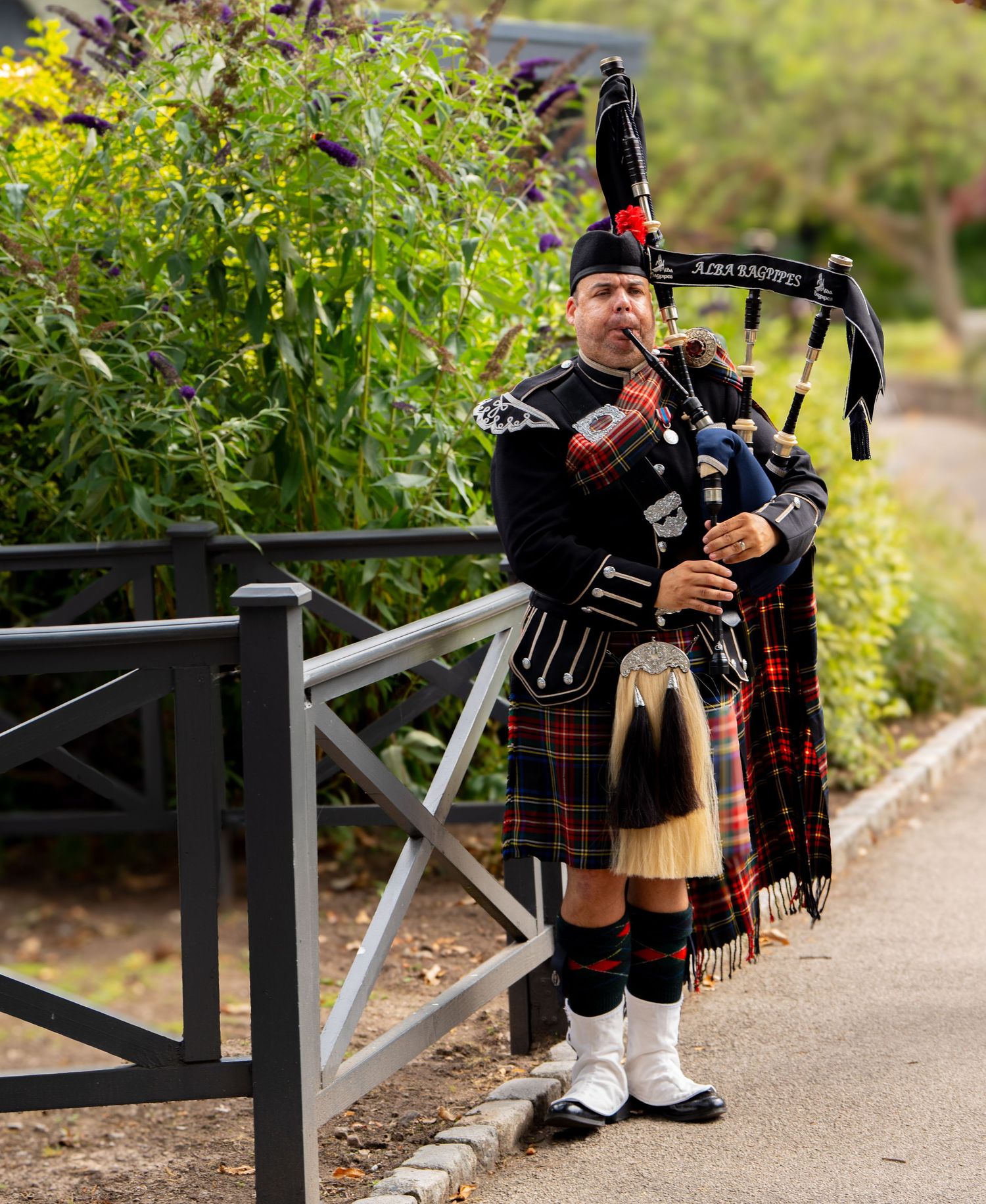 Scottish bagpiper in traditional tartan kilt and uniform performs outdoors at wedding.