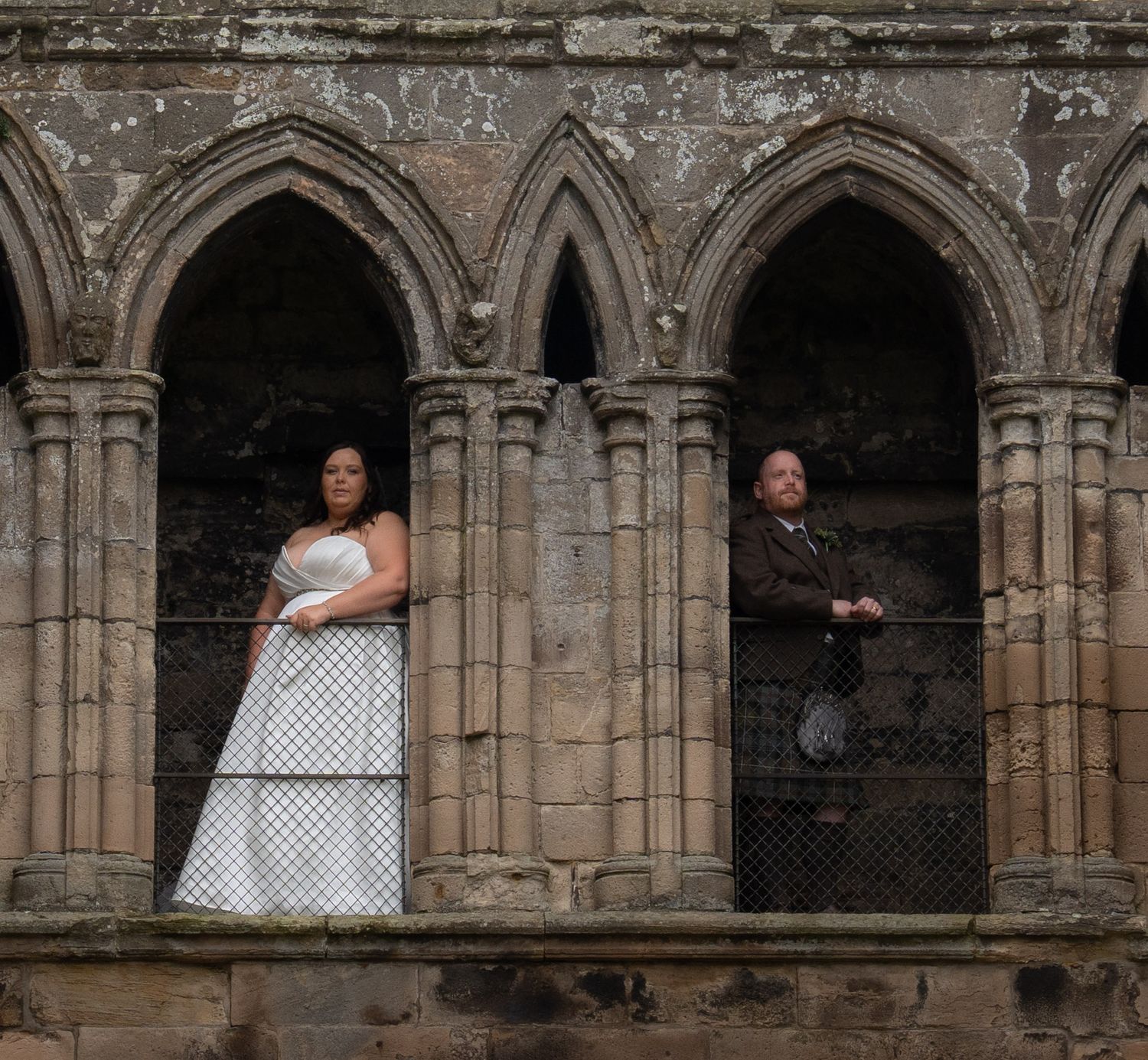 Bride and groom pose in Gothic stone arched windows of a historic ruined abbey.