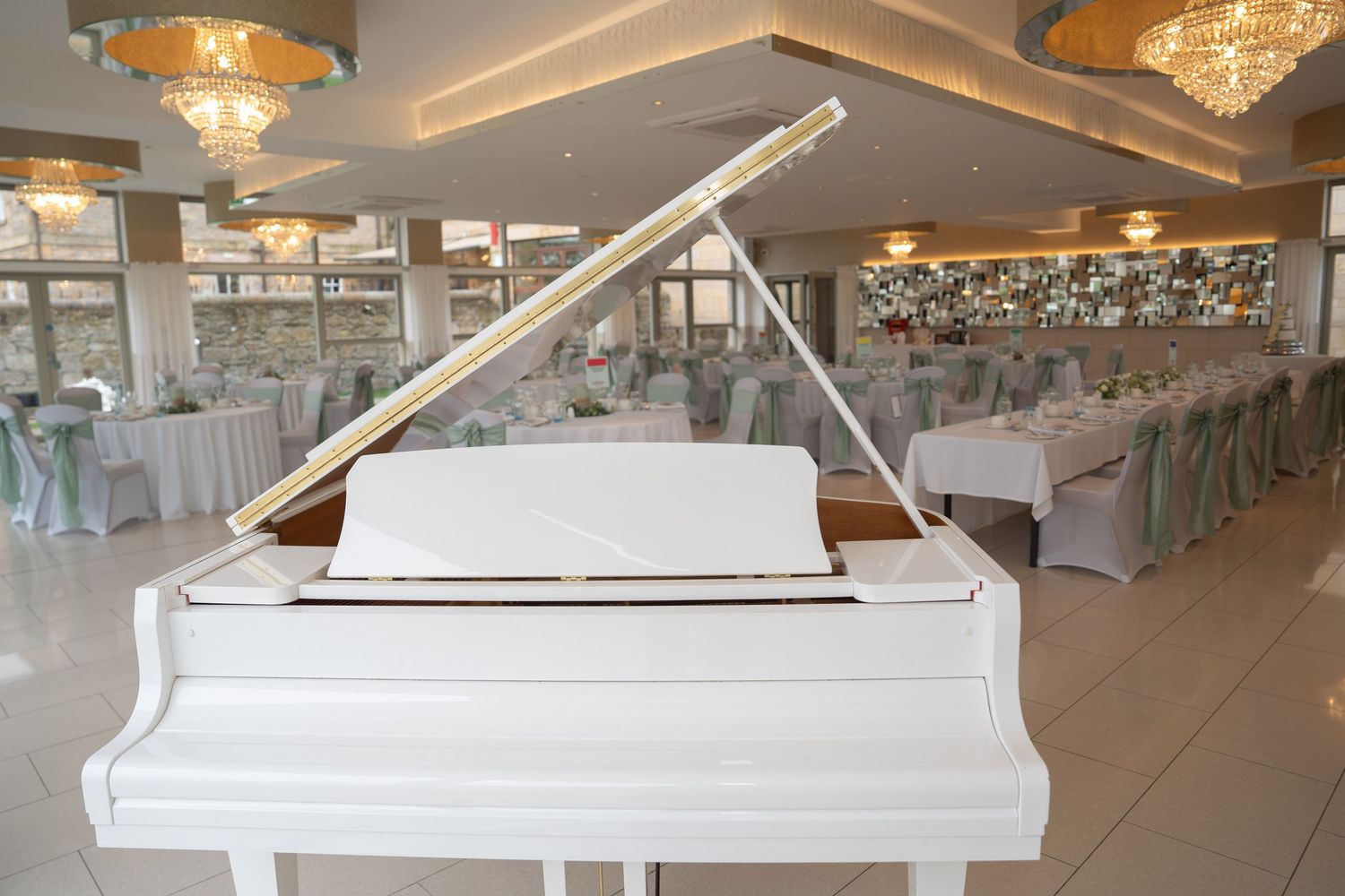 Elegant white grand piano in a decorated wedding reception venue with chandeliers and green chair covers.