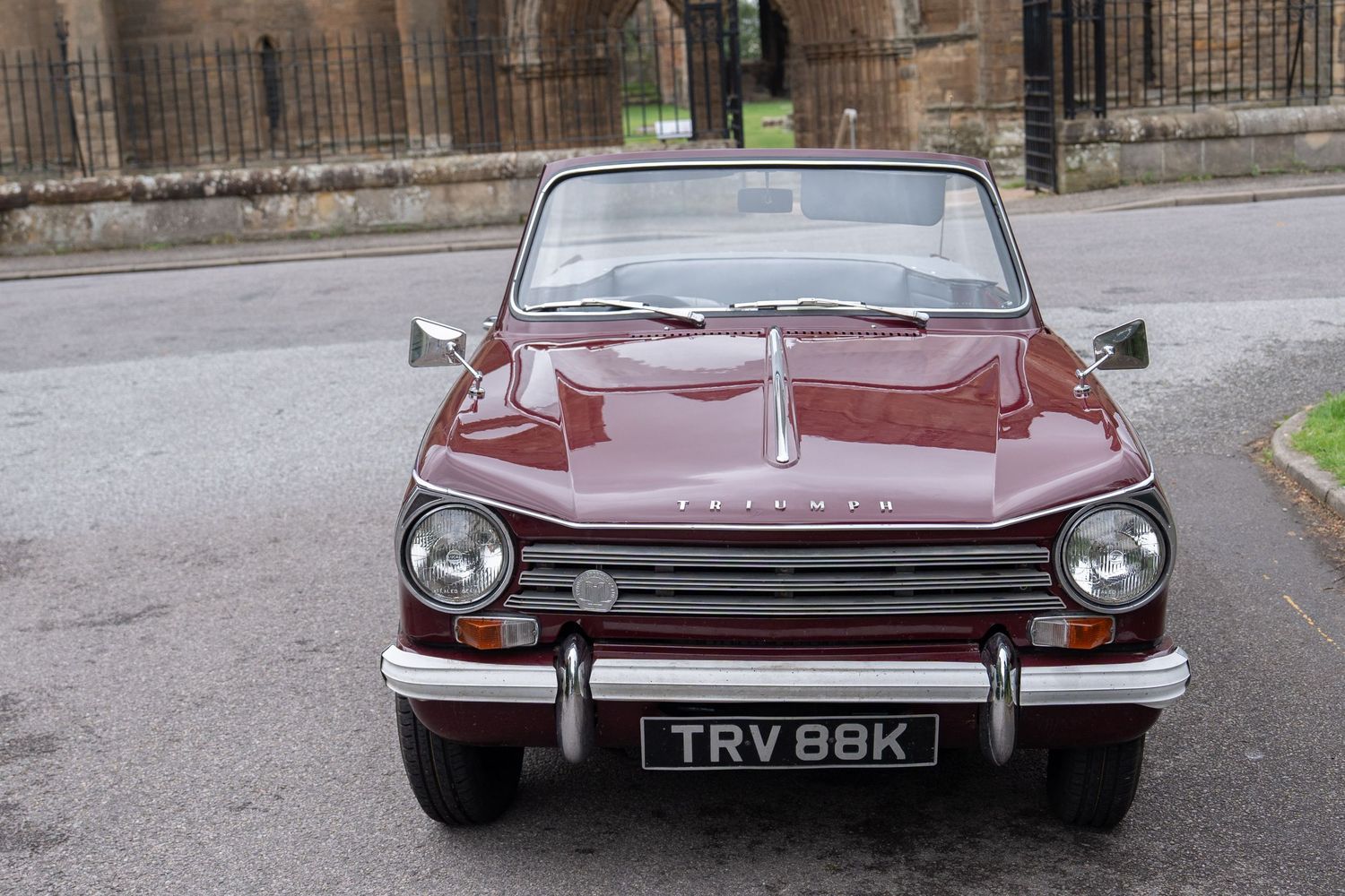 Vintage burgundy Triumph convertible classic car with registration TRV 88K parked outside a stone church building.