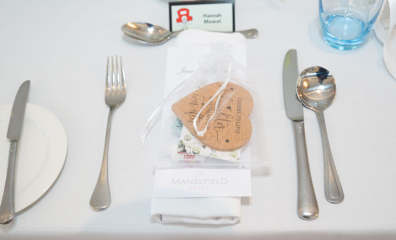 Elegant wedding table place setting with cutlery, name card, and heart-shaped cork favor on white linen tablecloth.