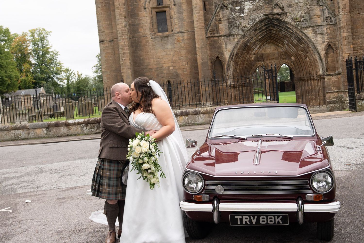 Newlyweds sharing a kiss beside a classic burgundy Triumph car outside a ruined Scottish stone cathedral.