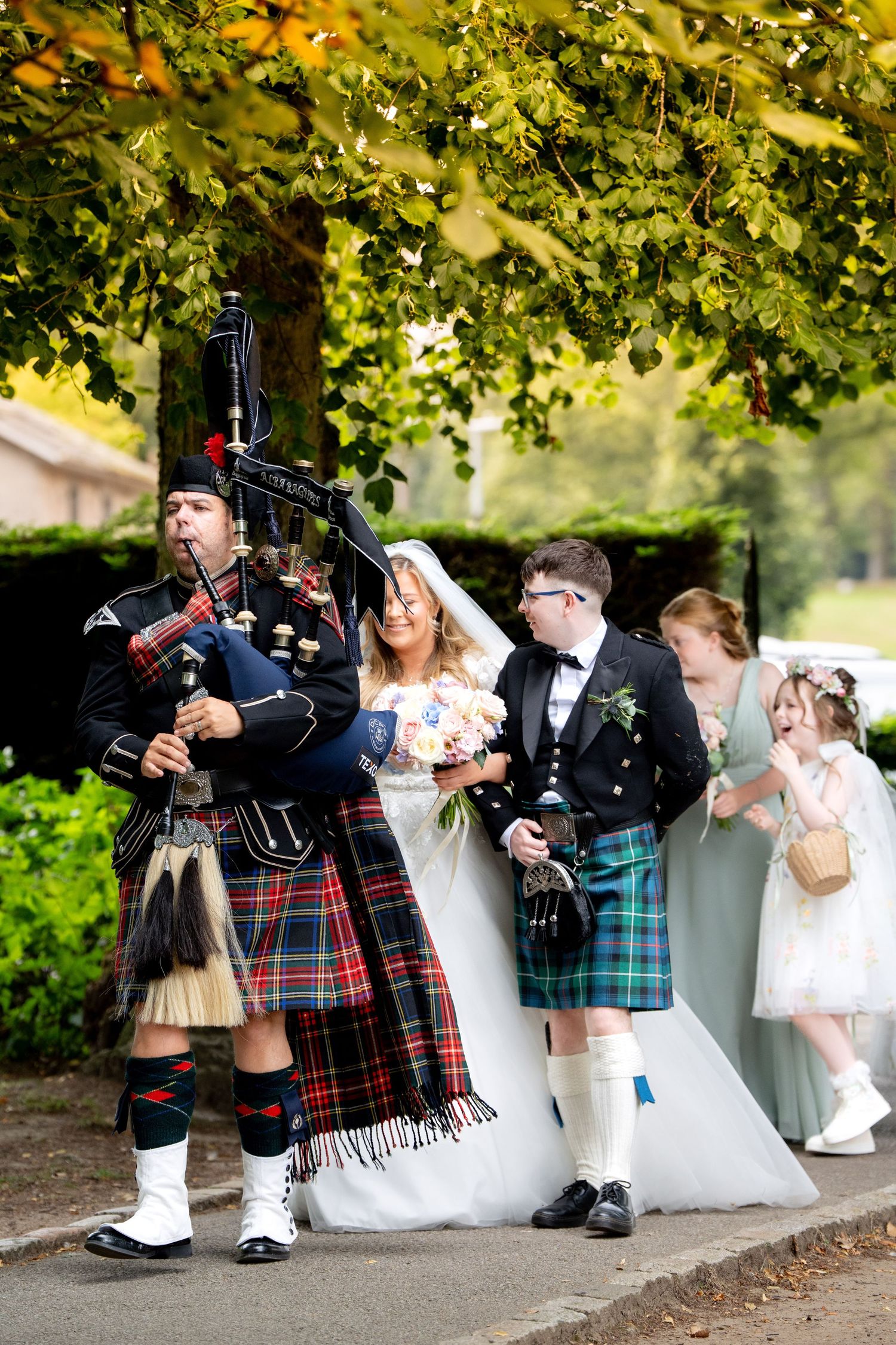 Scottish wedding procession with bagpiper in red tartan kilt leading bride and groom outdoors.
