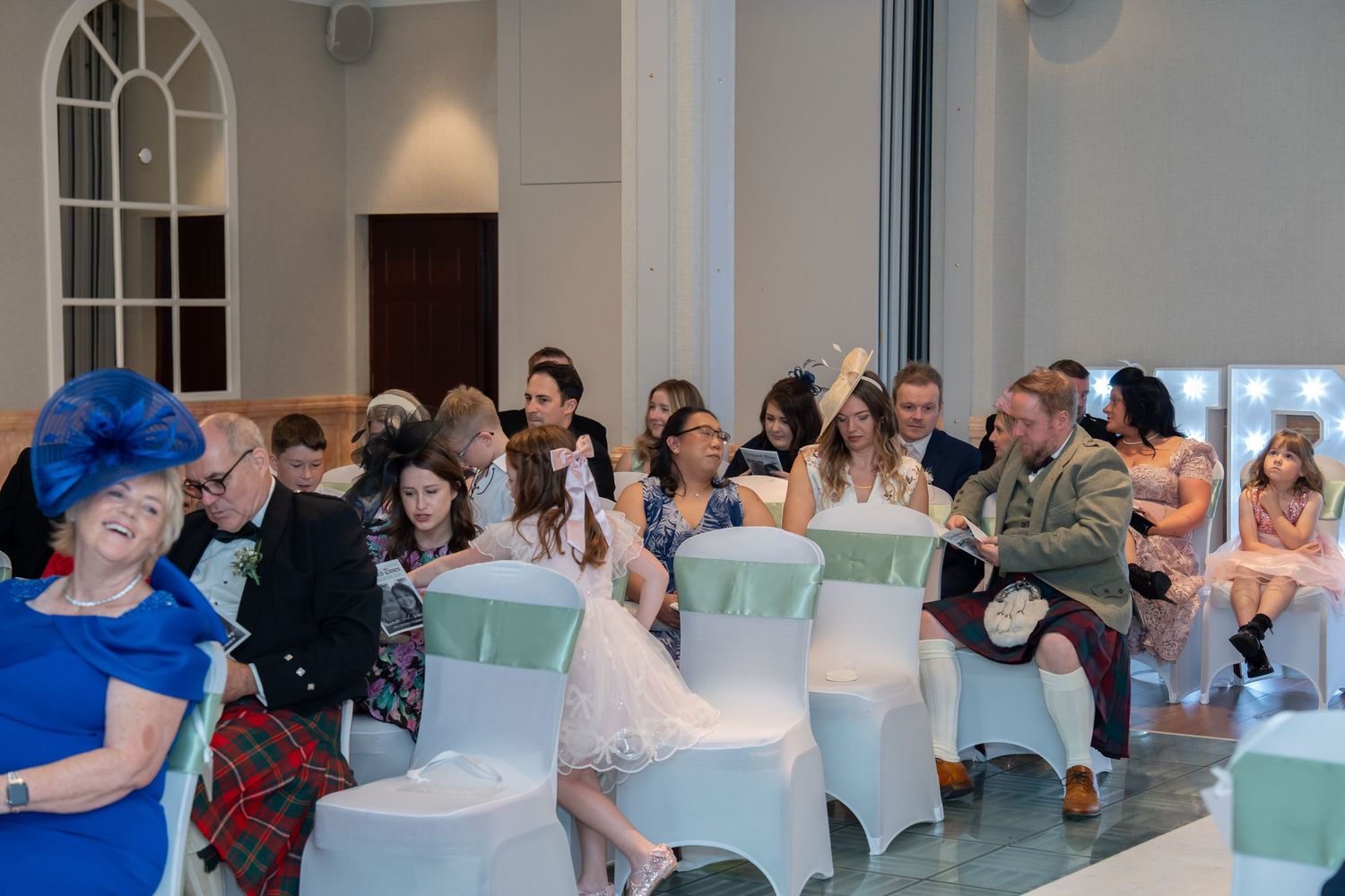 Wedding guests seated in white chair covers during a ceremony in a bright modern venue with tall arched windows.