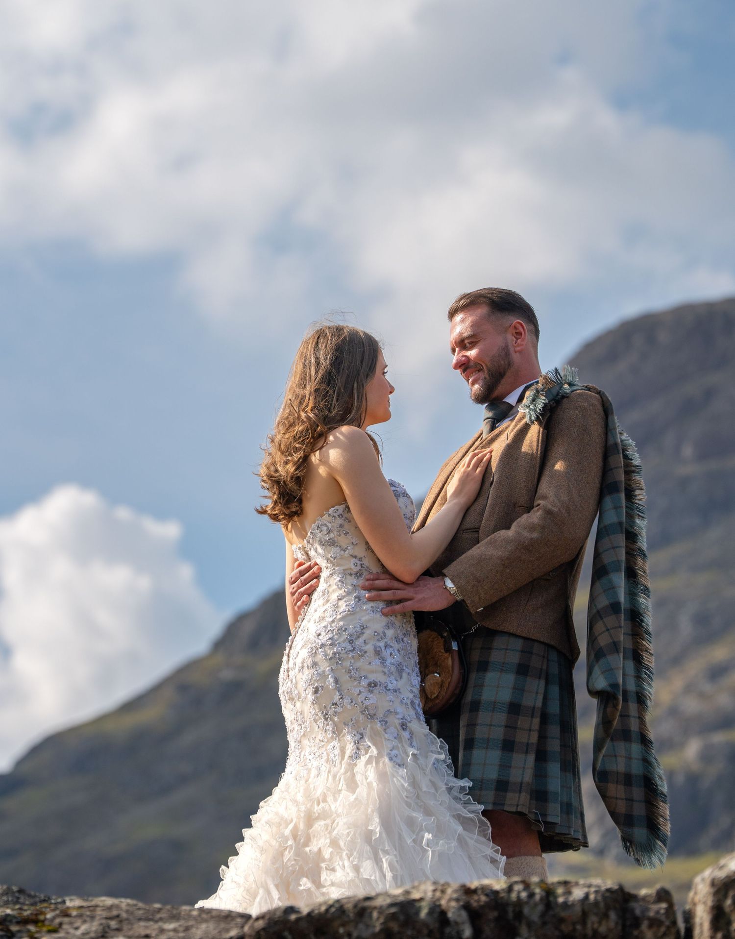 Bride in white mermaid gown and groom in tartan kilt embrace outdoors with Scottish Highland mountains behind them.