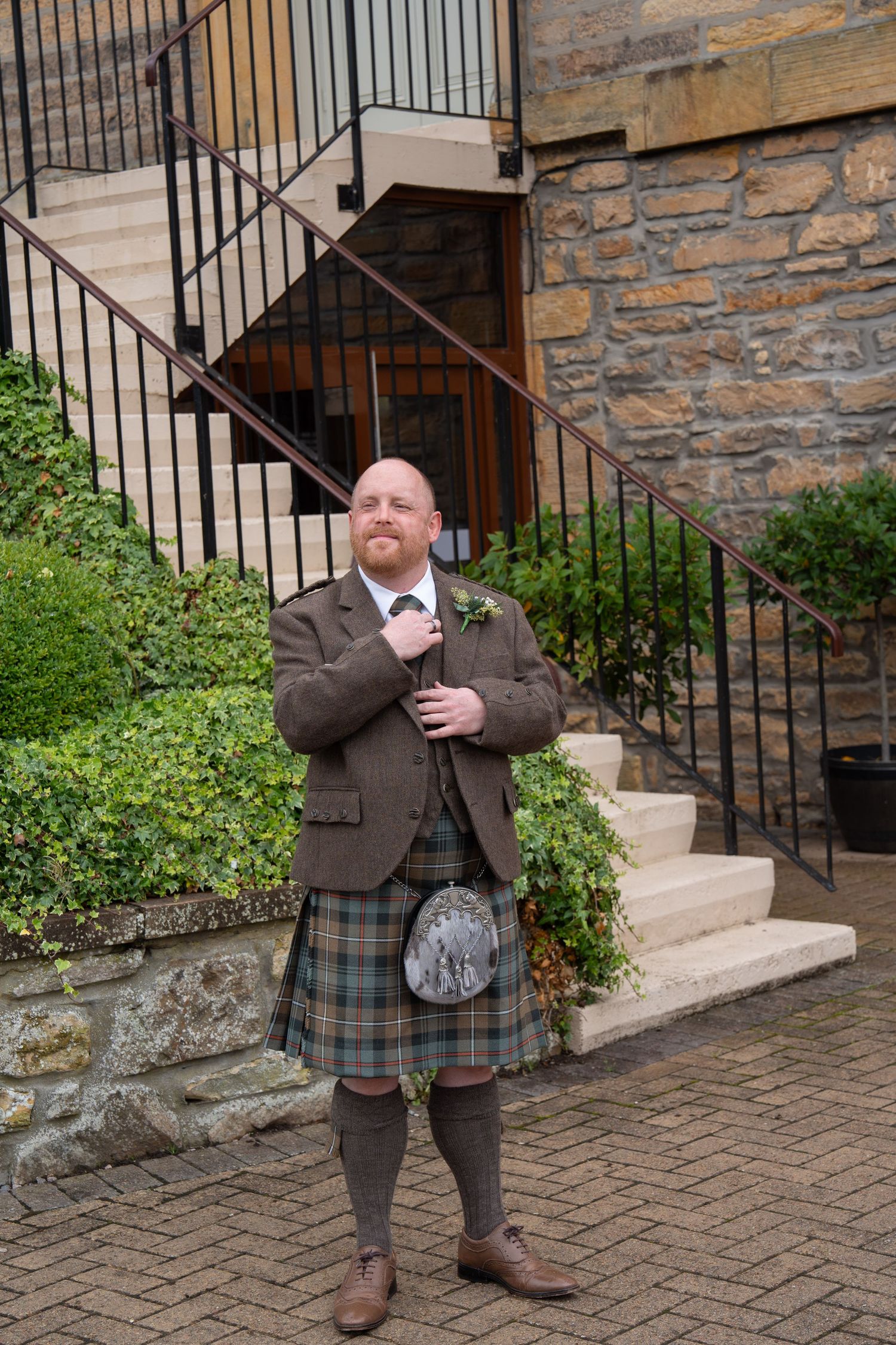 Groom in traditional Scottish tartan kilt, sporran, and tweed jacket standing outside stone building with staircase.
