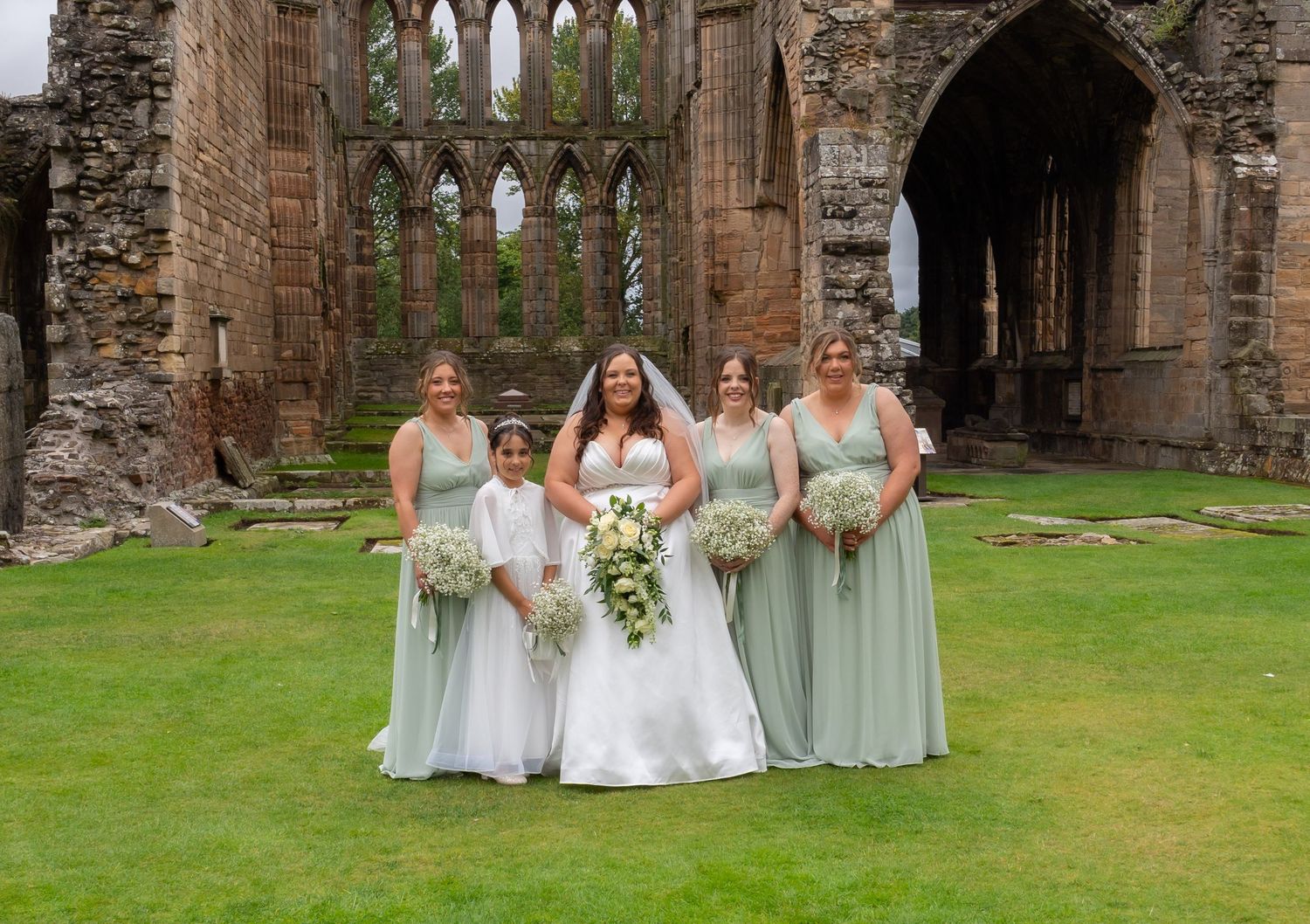 Bride and four bridesmaids in sage green dresses posing at a historic stone abbey ruins wedding venue.