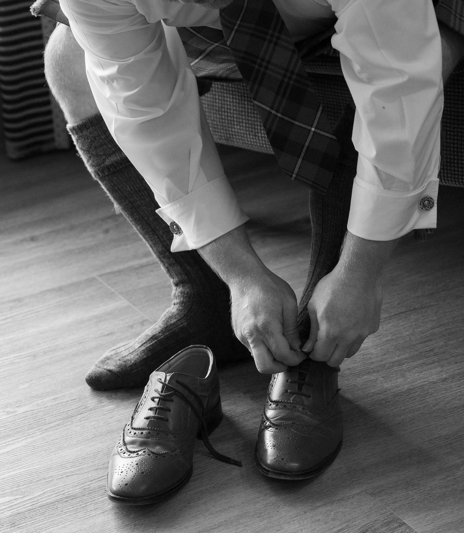 Black and white photo of groom in tartan kilt lacing up traditional Scottish brogue shoes on wooden floor.