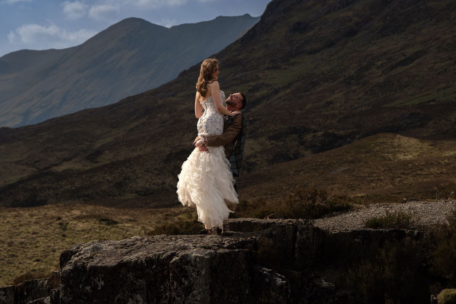 Groom lifting bride in white ruffled dress on rocky outcrop with dramatic Scottish Highland mountains backdrop.