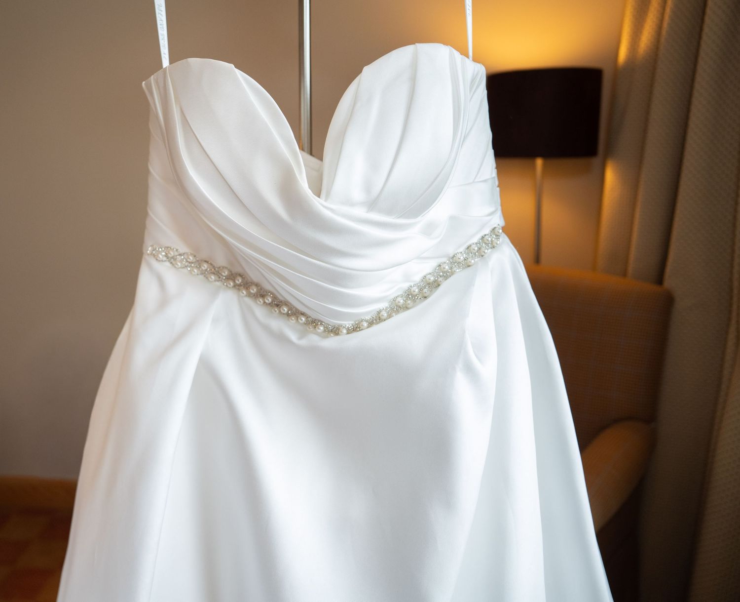 Elegant strapless white wedding dress with pearl and crystal embellished waistband hanging in a warmly lit hotel room.