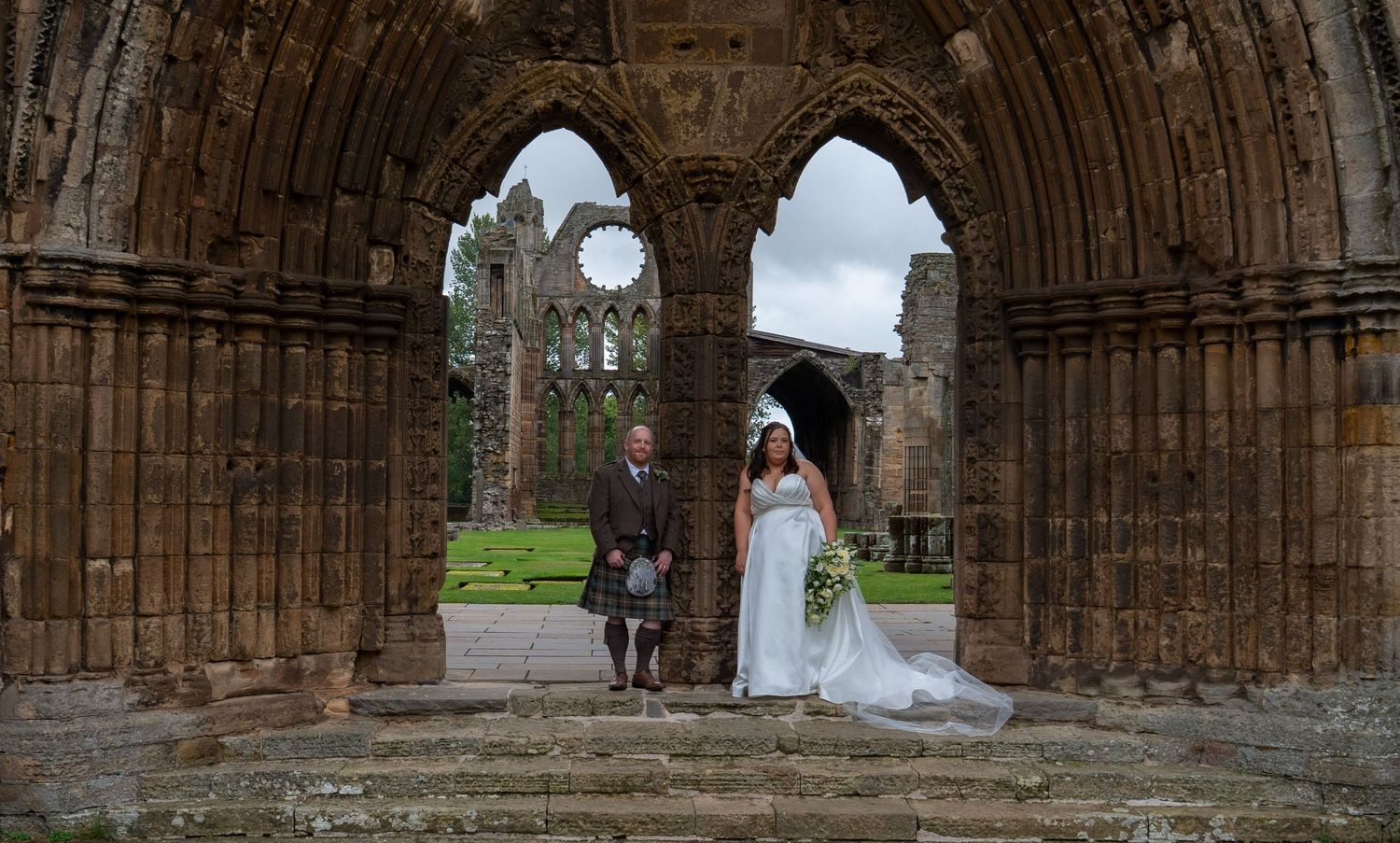 Bride and groom posing inside the ancient stone arches of a Scottish abbey ruins.
