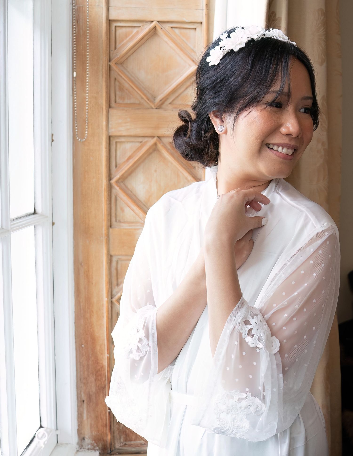 A smiling bride in a white lace robe and floral headpiece stands by a window.