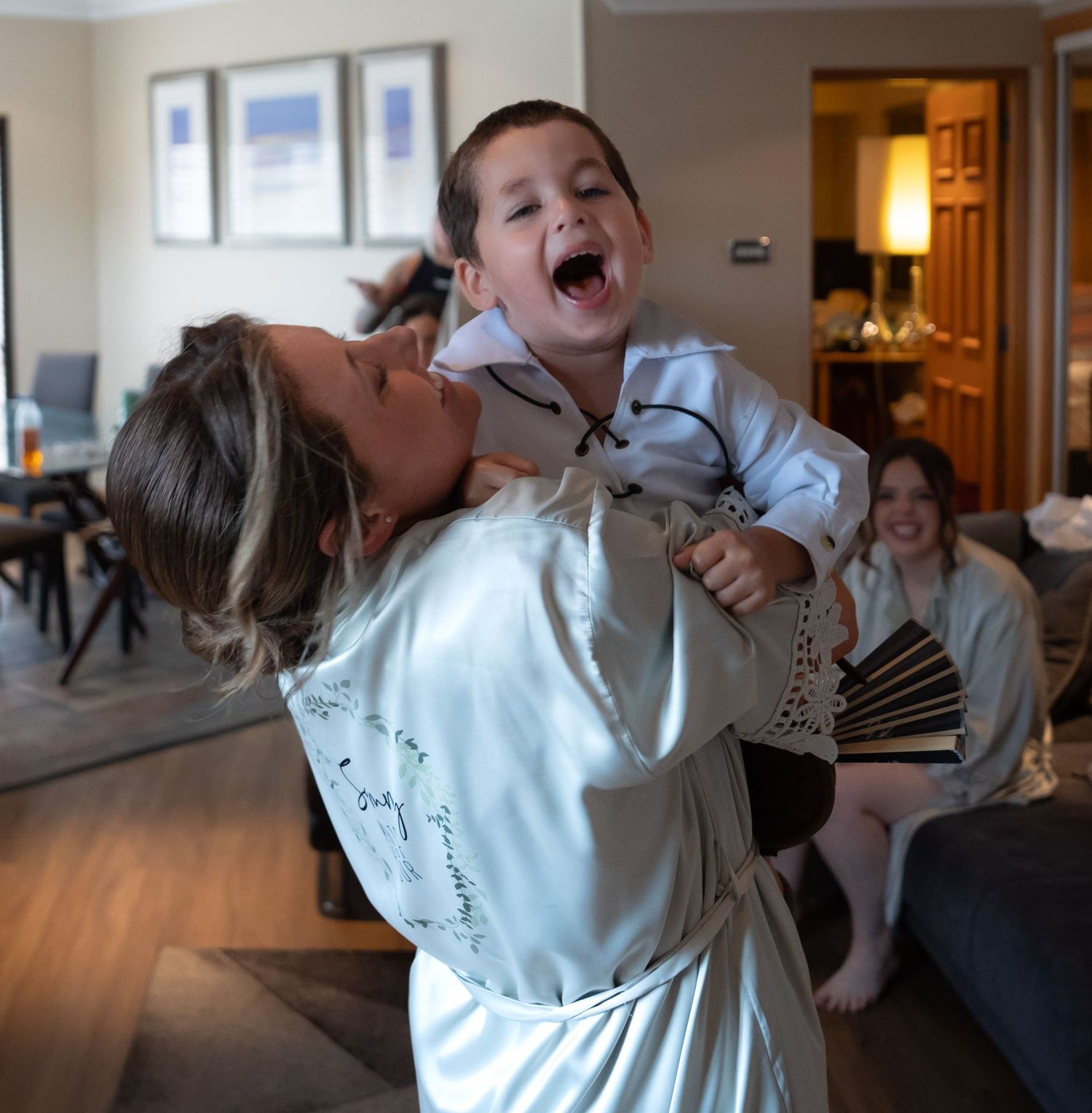 Joyful woman in bridal robe lifting laughing young boy dressed in formal attire during wedding preparations.