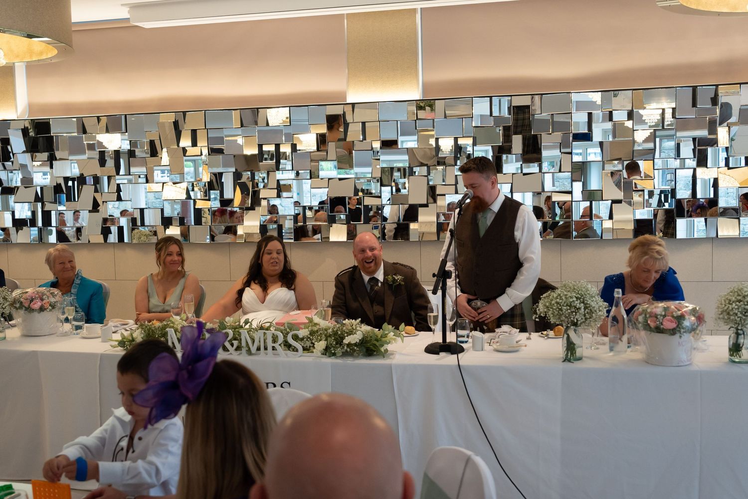 Groom giving wedding speech at top table with bride and wedding party seated before mirrored wall backdrop.