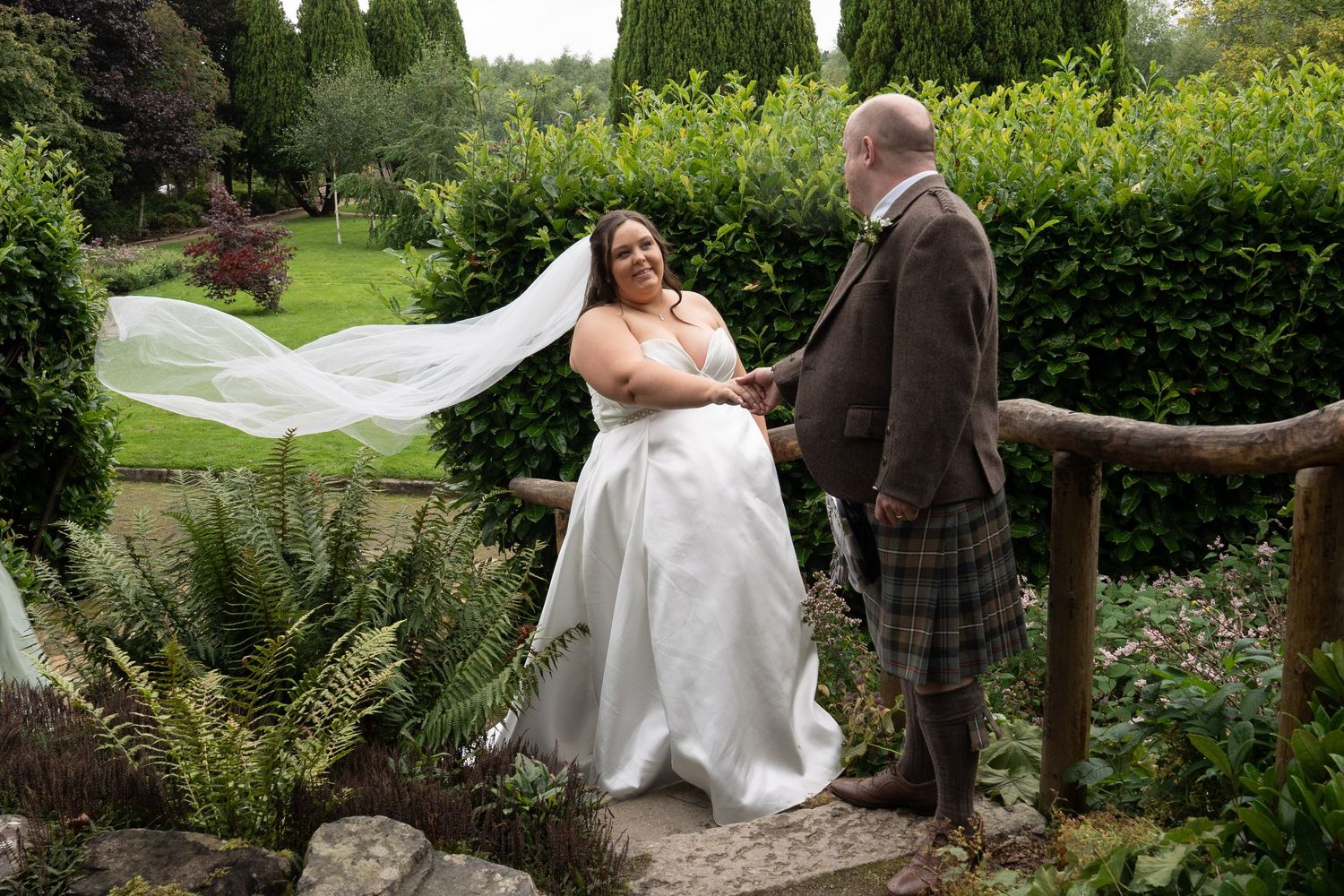 Bride in white gown with flowing veil stands with groom in tartan kilt in a lush garden setting.