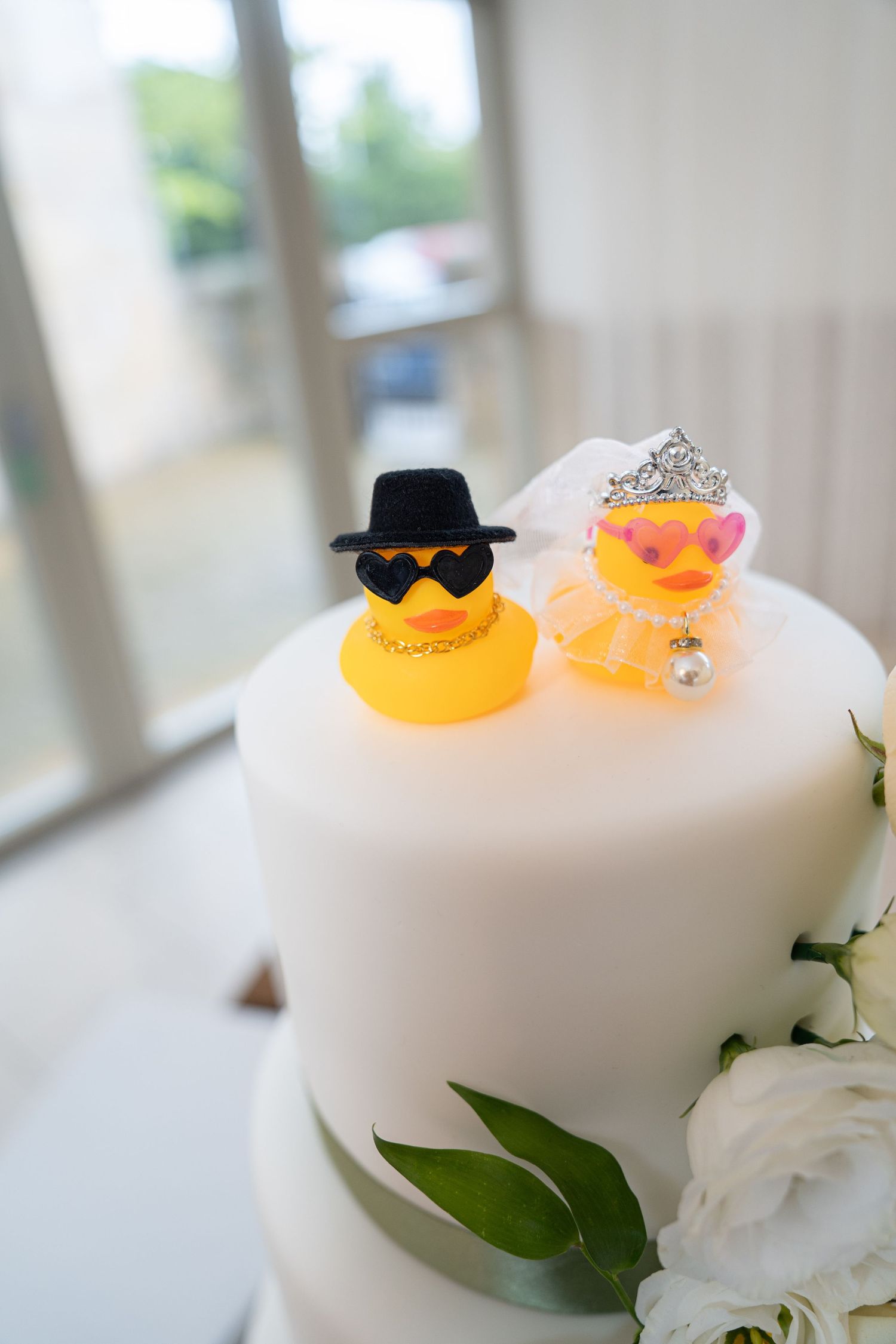 White wedding cake topped with bride and groom rubber ducks wearing tiara and top hat decorations.