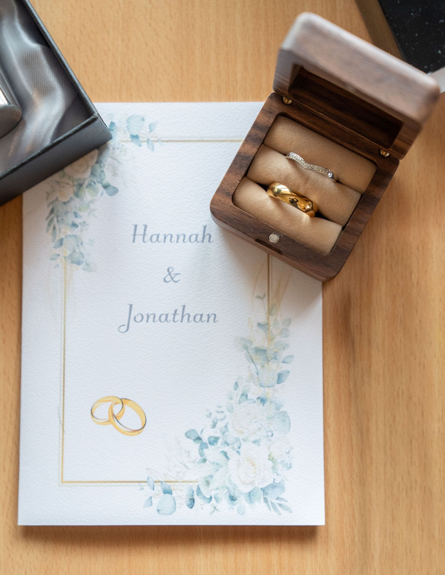 Wedding stationery flat lay featuring Hannah and Jonathan order of service with gold rings and wooden ring box.