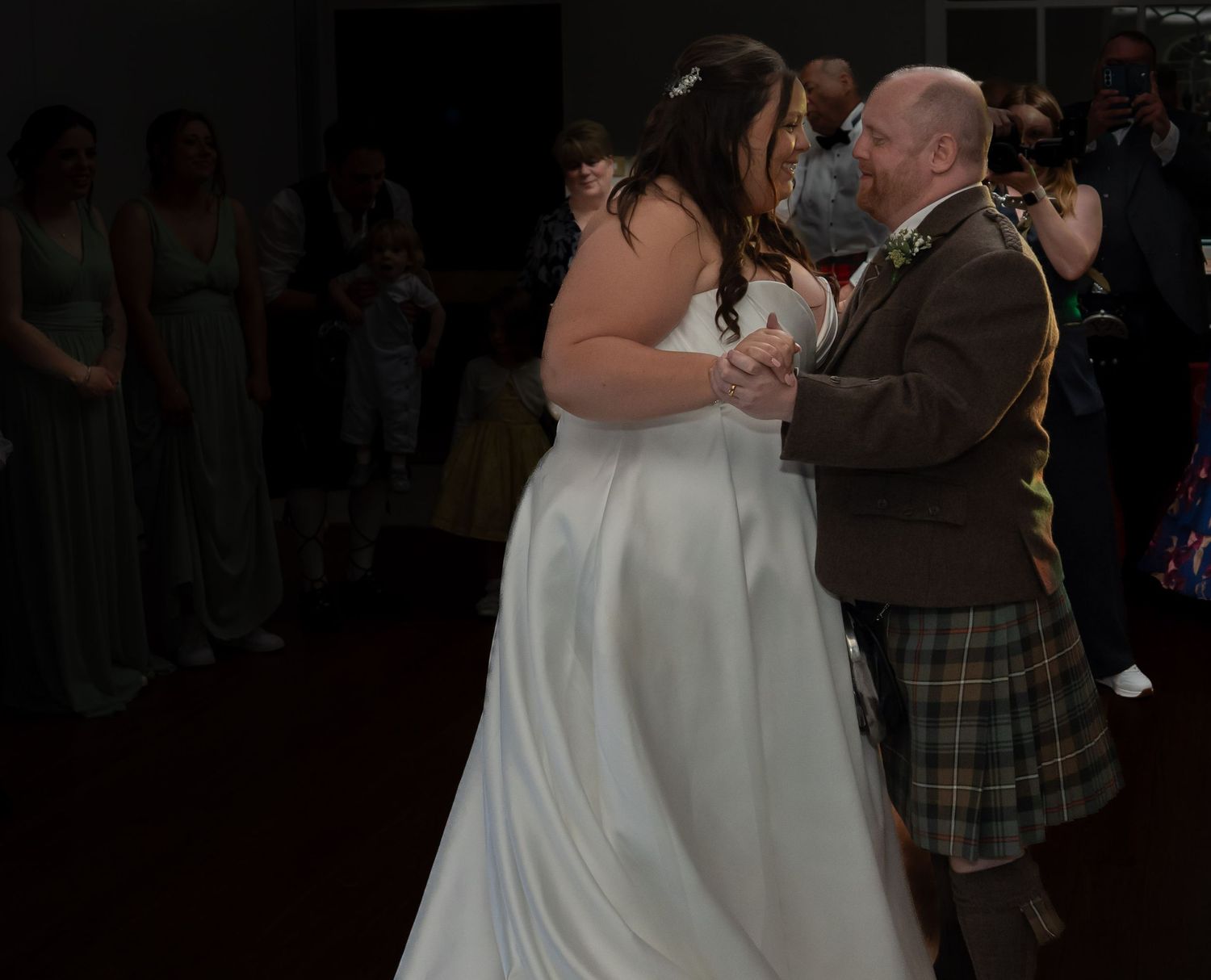 Bride in white satin gown shares first dance with groom in traditional Scottish tartan kilt at wedding reception.