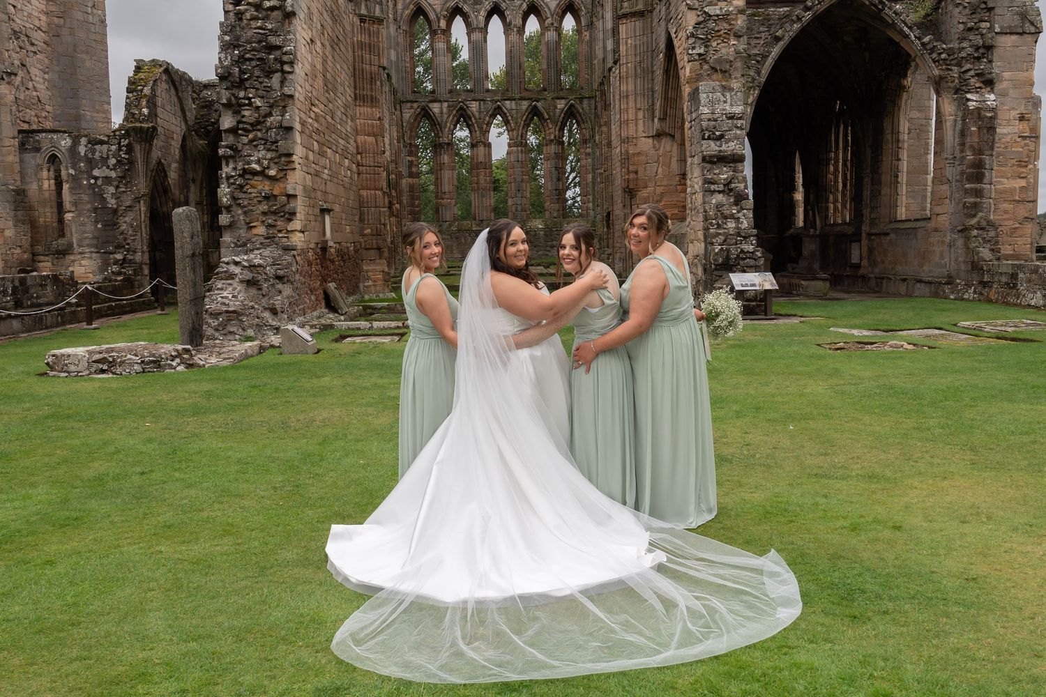 Bride in white gown with long veil poses with three bridesmaids in sage green dresses at historic abbey ruins.