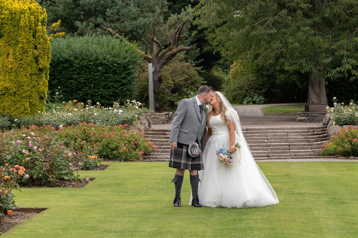 Bride and groom sharing a romantic moment in a lush garden, groom wearing a traditional Scottish kilt.