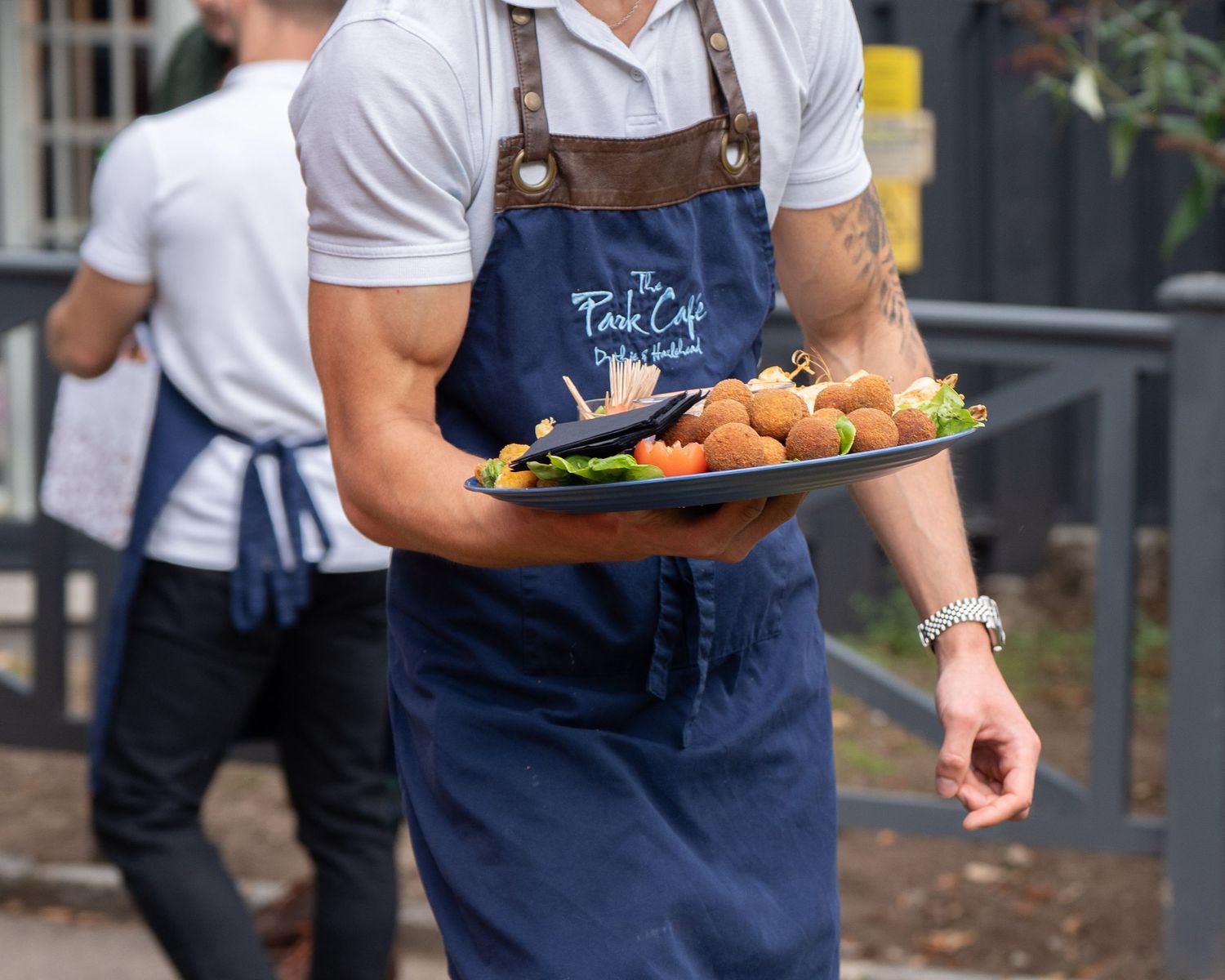 Catering staff member in blue apron carries platter of fried appetizers at outdoor event.