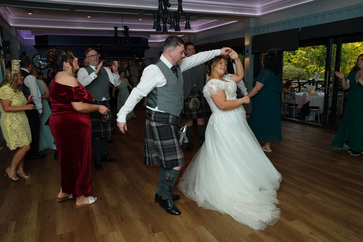 Bride in white gown and groom in Scottish kilt enjoy a lively dance with wedding guests on the dance floor.