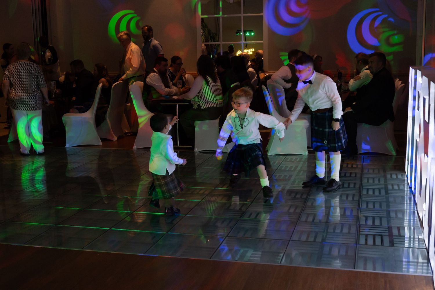 Two young boys in kilts dancing energetically on a lit dance floor at a wedding reception.